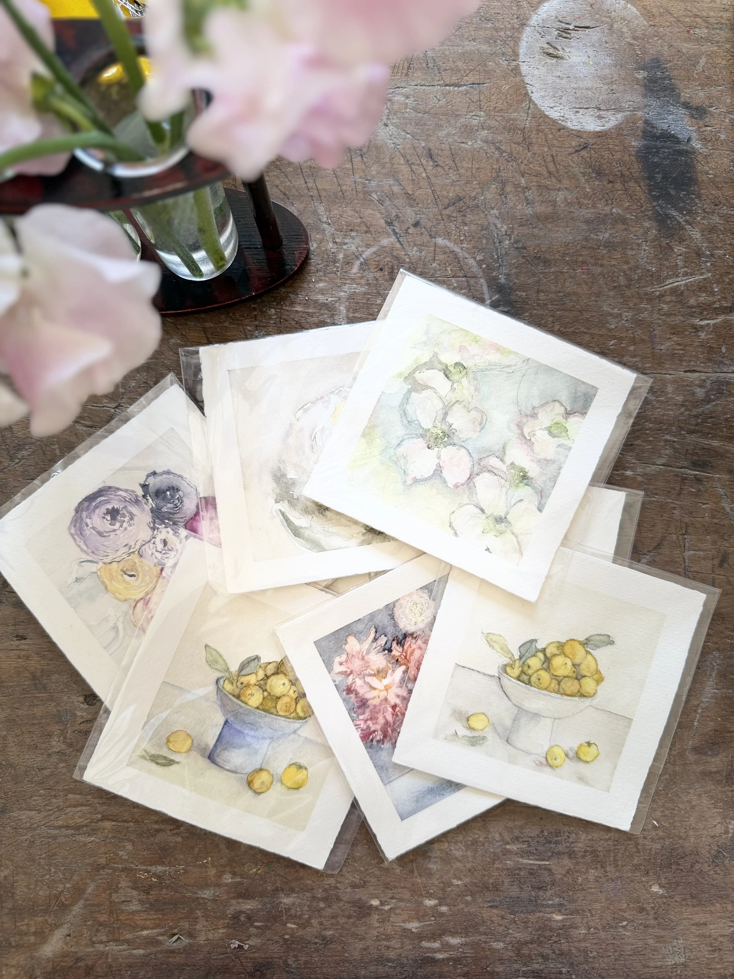 A collection of small watercolor paintings of flowers and fruit arranged on a wooden table, with a vase of pink flowers in the top left corner.