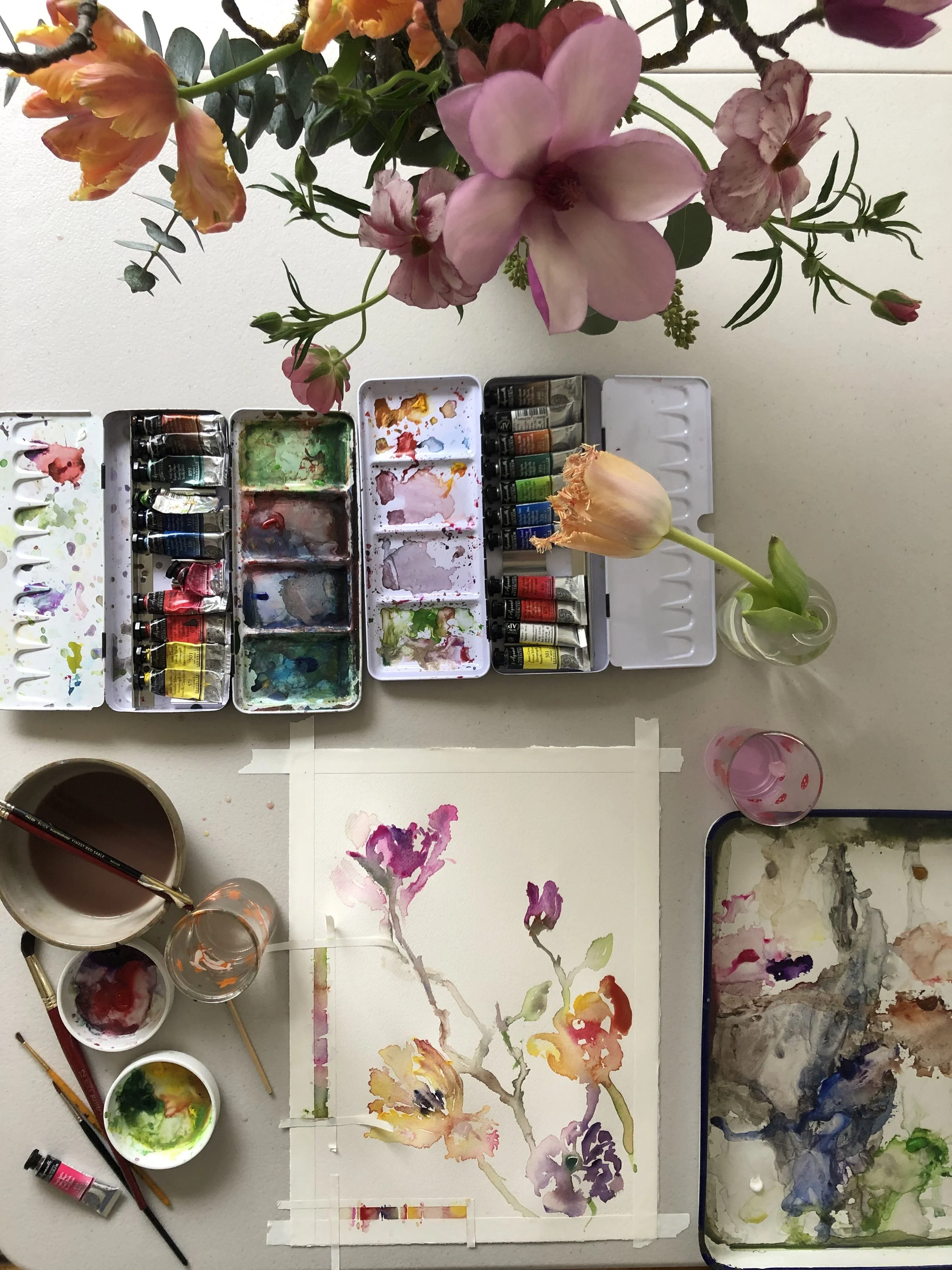 A watercolor painting of a flower on paper, surrounded by various watercolor paints, brushes, and containers with water. There is a vase with a single pink tulip and a larger bouquet of pink and orange flowers above.