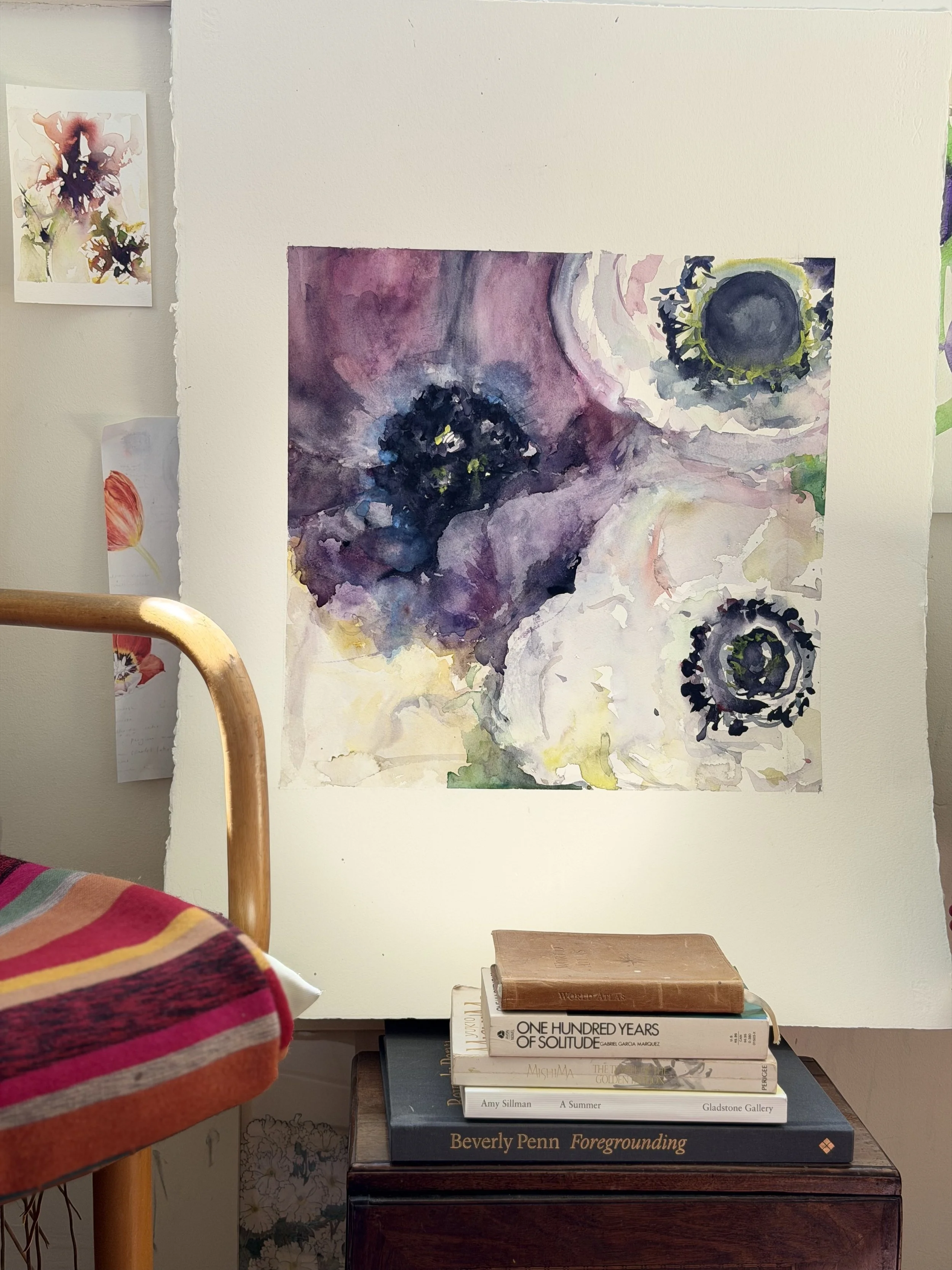 An abstract watercolor painting of flowers on a white wall, with a stack of art books below it, including titles such as 'One Hundred Years of Solitude' and 'Foregrounding.'