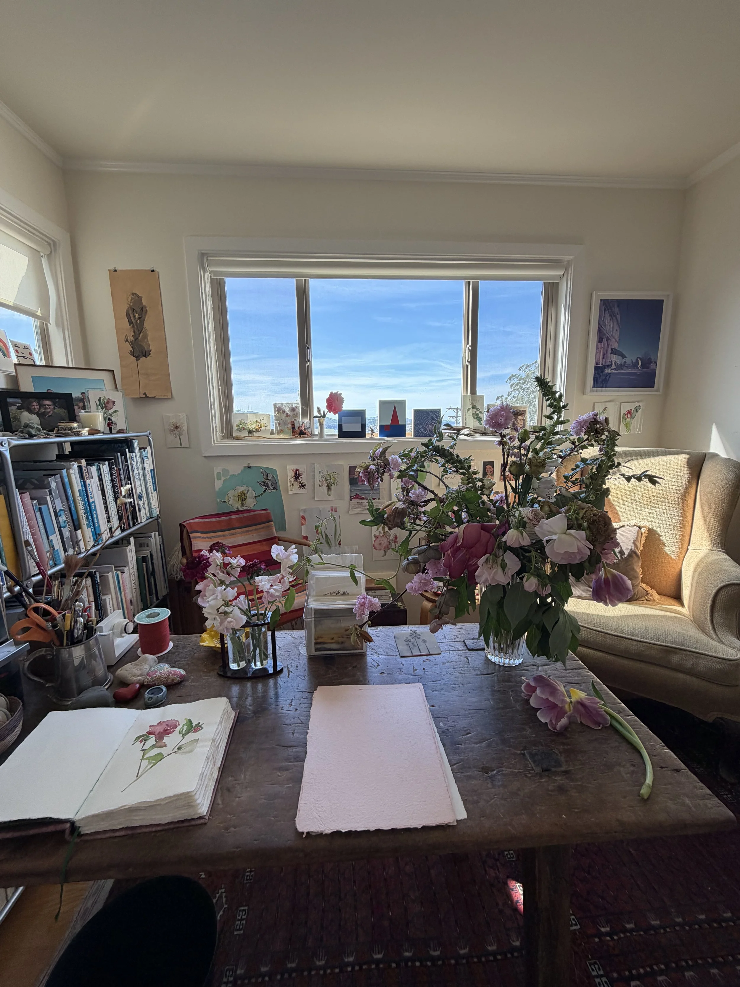 Interior of a room with a wooden table holding flowers in a glass vase, artwork, and books. There is a window with a view of a blue sky. A beige sofa and a bookshelf are visible in the background.
