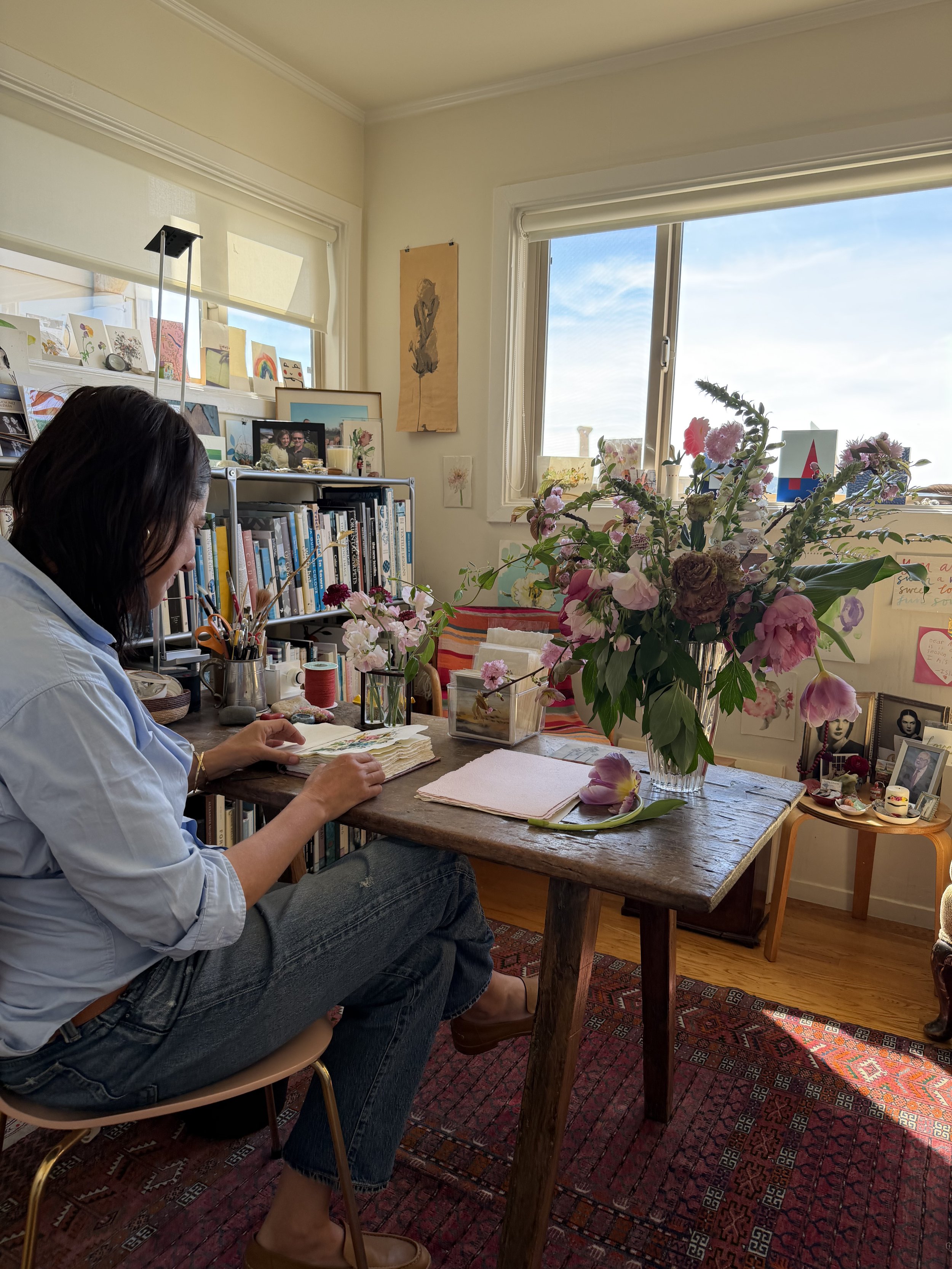 A woman sitting at a wooden table with a large flower arrangement, working in a bright, decorated room with books, artwork, and sunlight through a window.