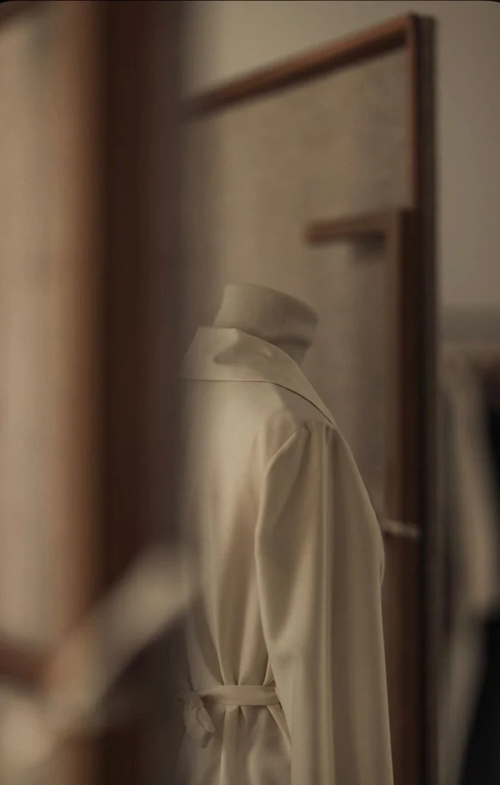 A cream-colored robe hanging on a wooden hanger, reflected in a slightly blurry mirror.