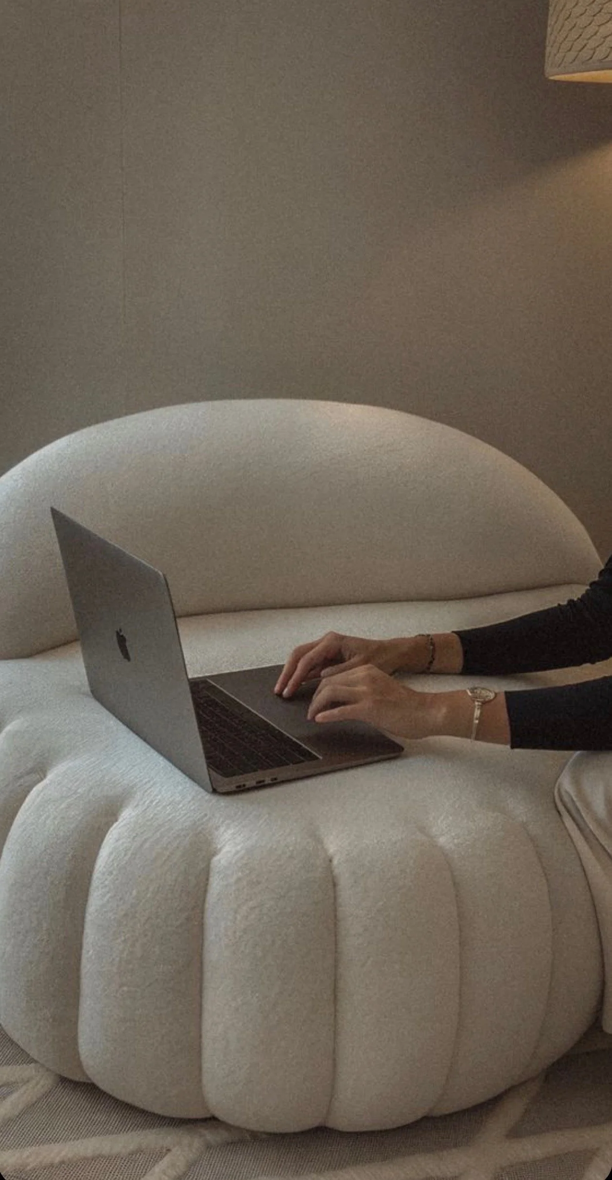 A person with long sleeves and a watch on their left wrist typing on a silver MacBook laptop, which is resting on a soft, cream-colored, rounded ottoman in a cozy interior space.
