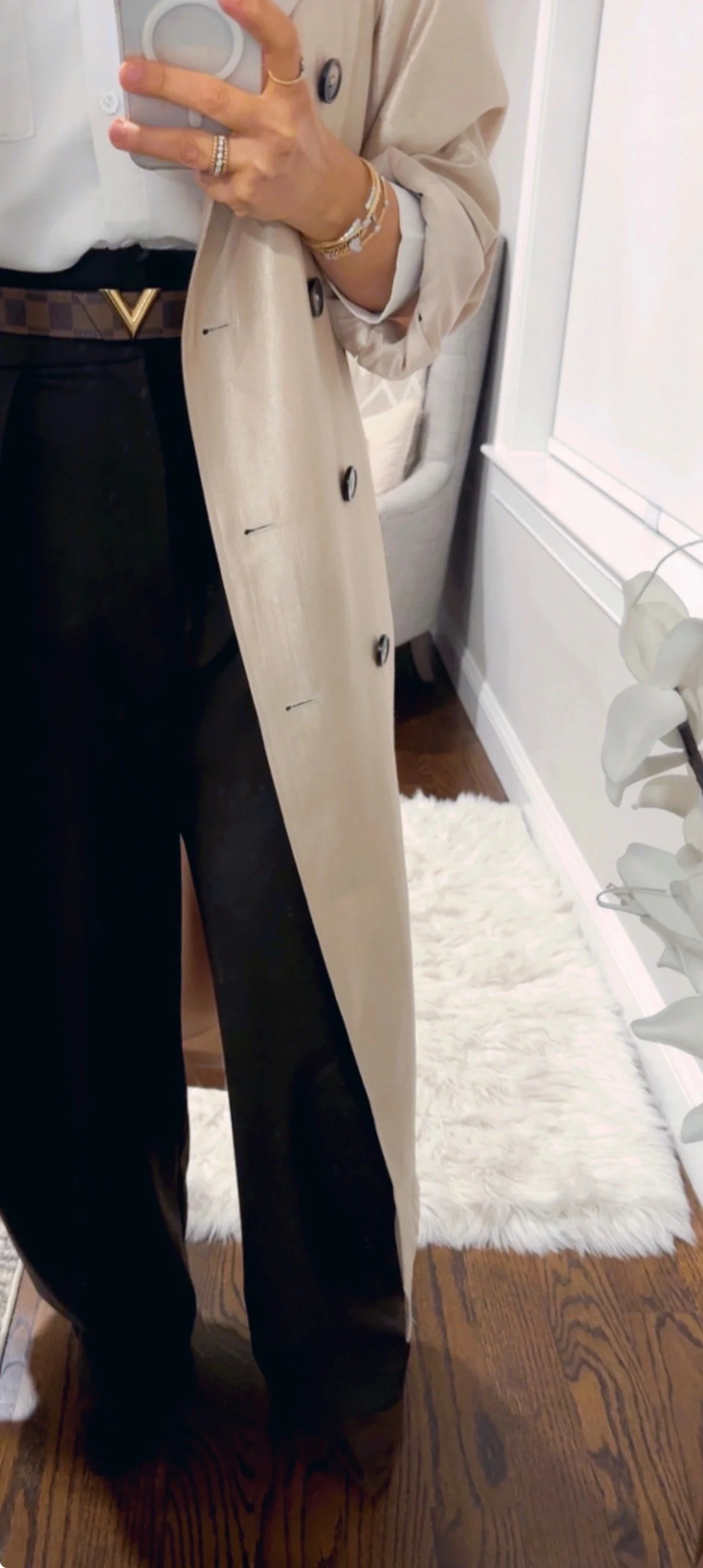 Mirror selfie of person holding a phone, wearing black high-waisted pants, a beige trench coat, visible rings, bracelets, and belt, standing in a room with hardwood flooring, white shag rug, and a window with sheer curtains.