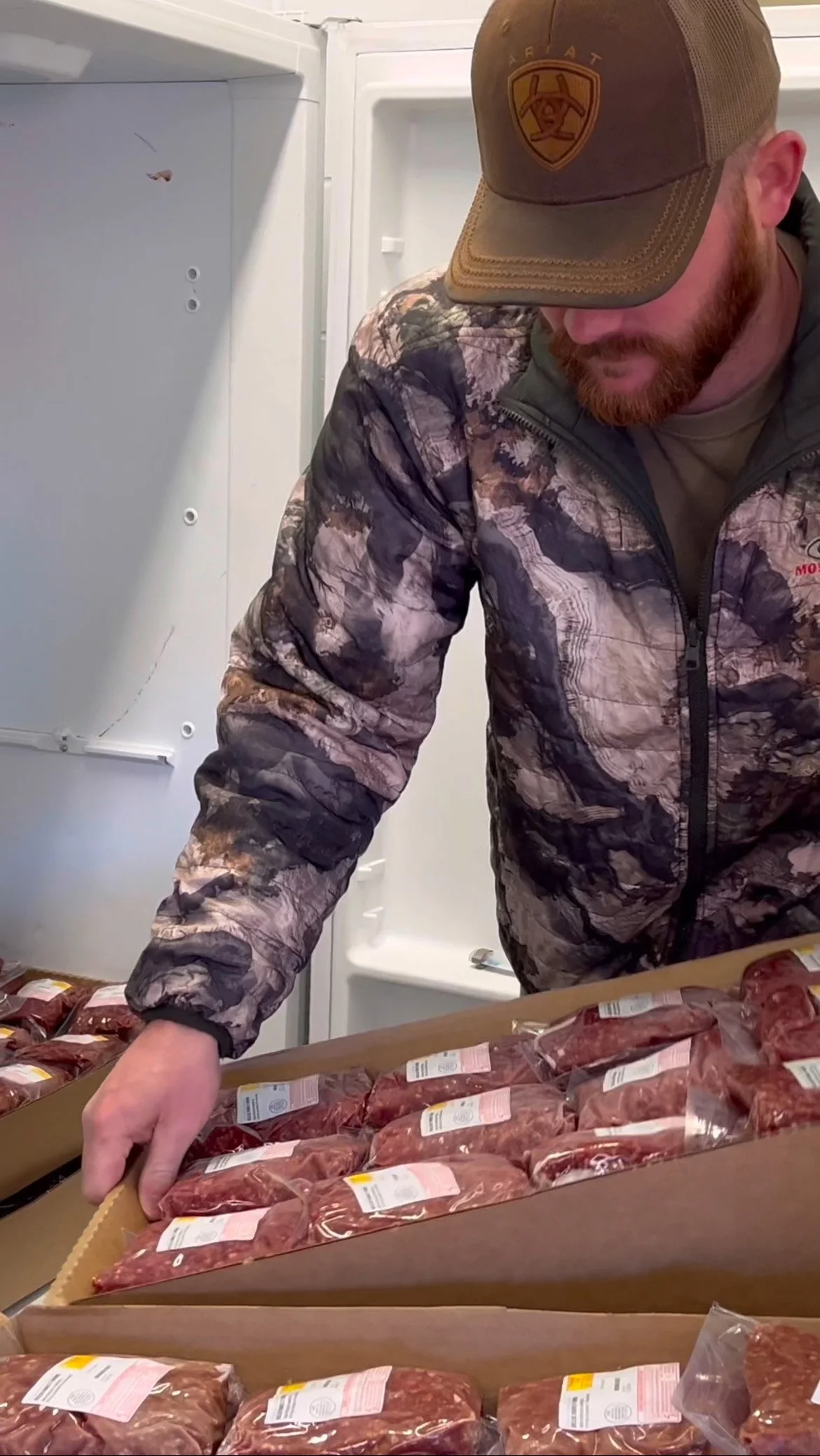 Man in camouflage jacket and brown cap with a badge inside a refrigerator, inspecting packages of raw meat on a table.