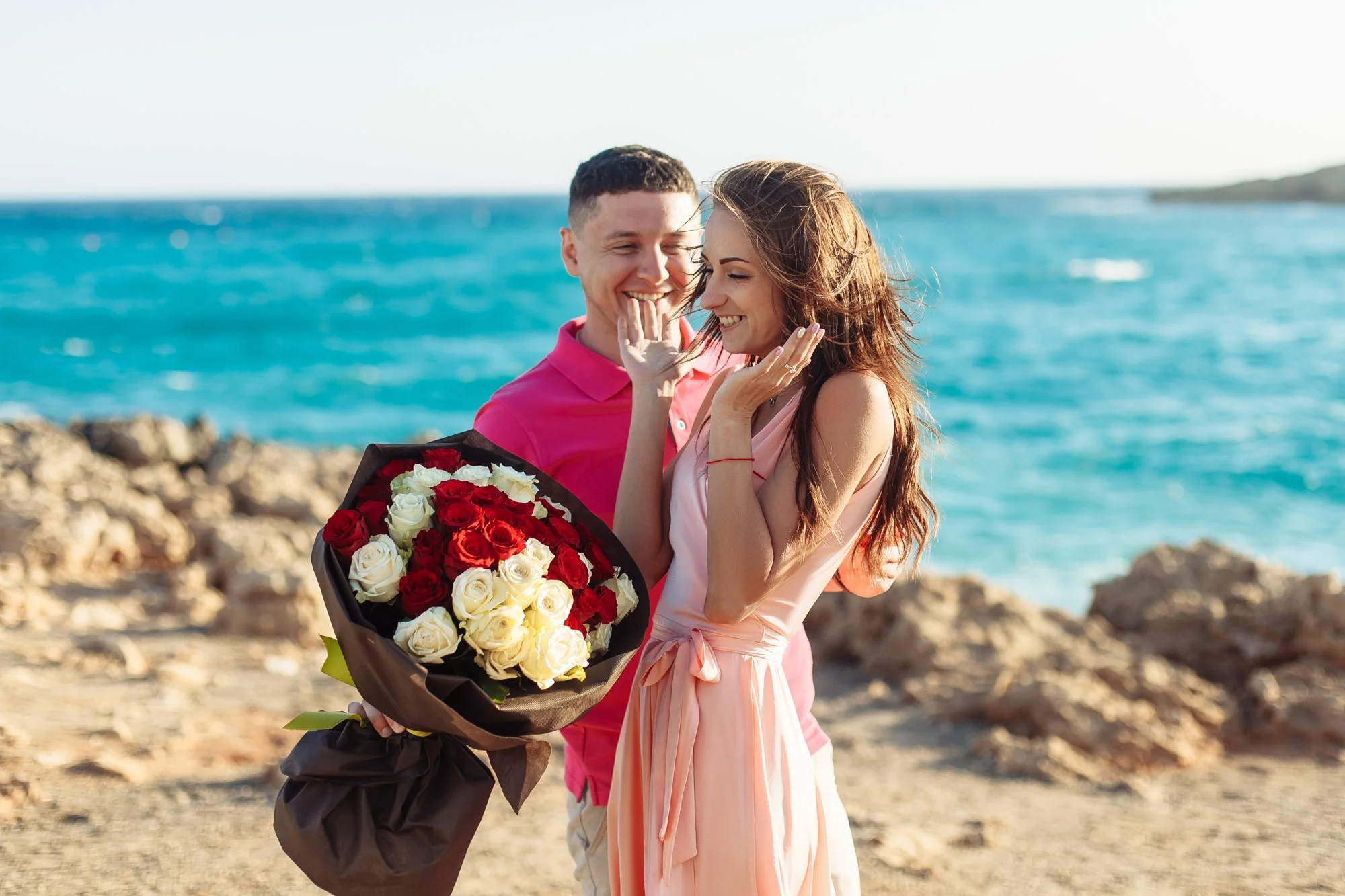 A man giving a woman a bouquet of red and white roses on a rocky beach with the ocean in the background in AYIA Napa
