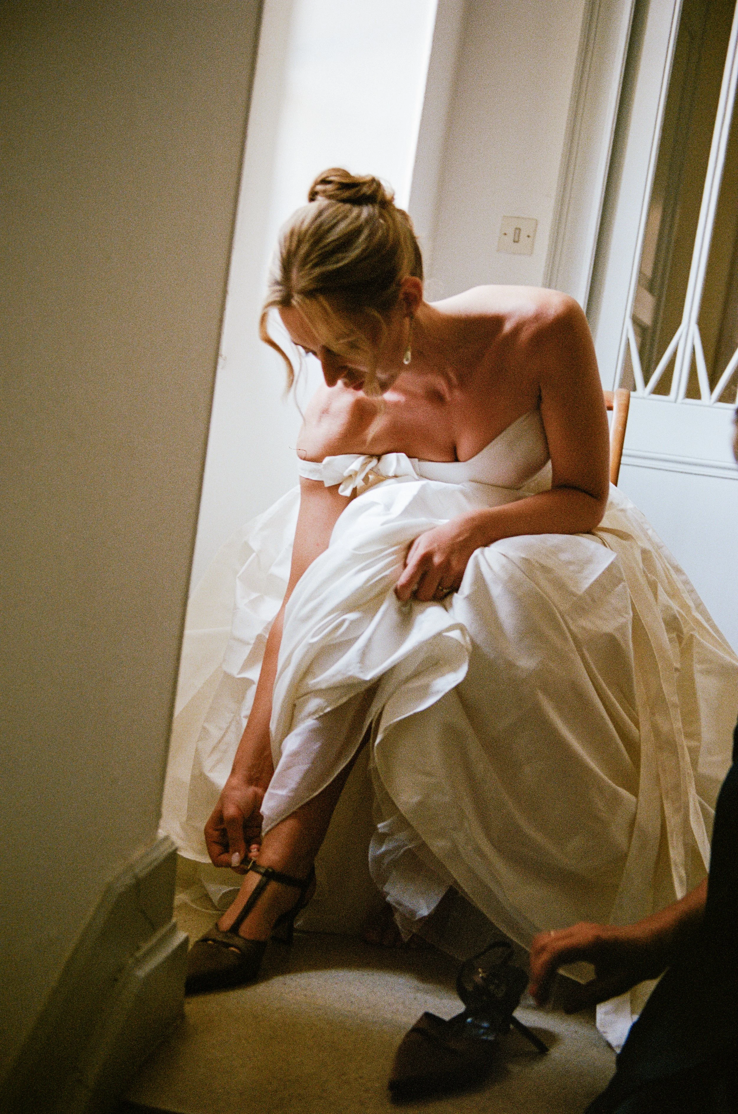 A woman dressed in a wedding gown adjusting her shoe while sitting on a chair in a small room, with someone else assisting or nearby.