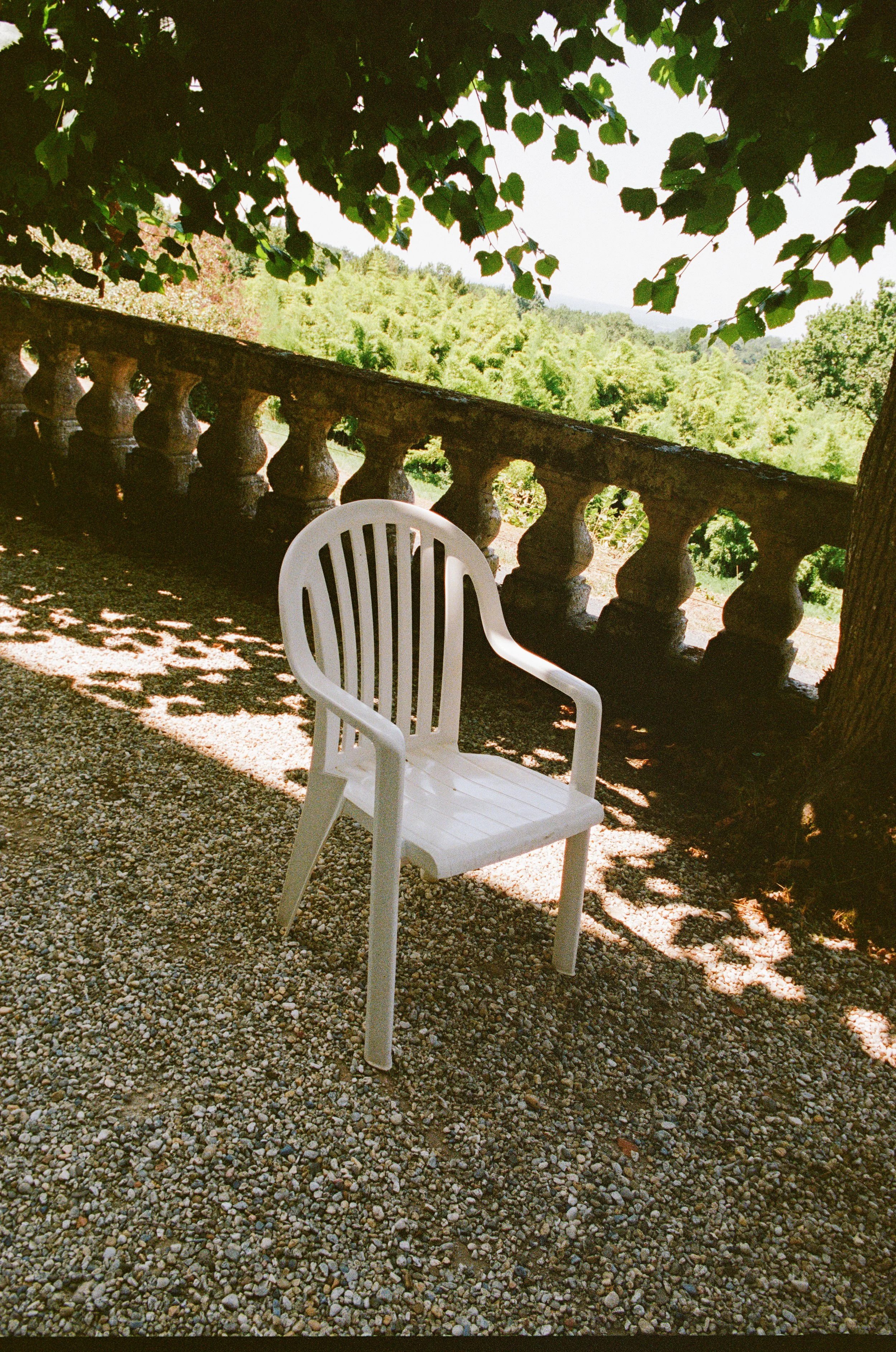 A white plastic chair on pebbled ground under a tree with green leaves. In the background, there is a stone balustrade and a view of lush green trees and hills.
