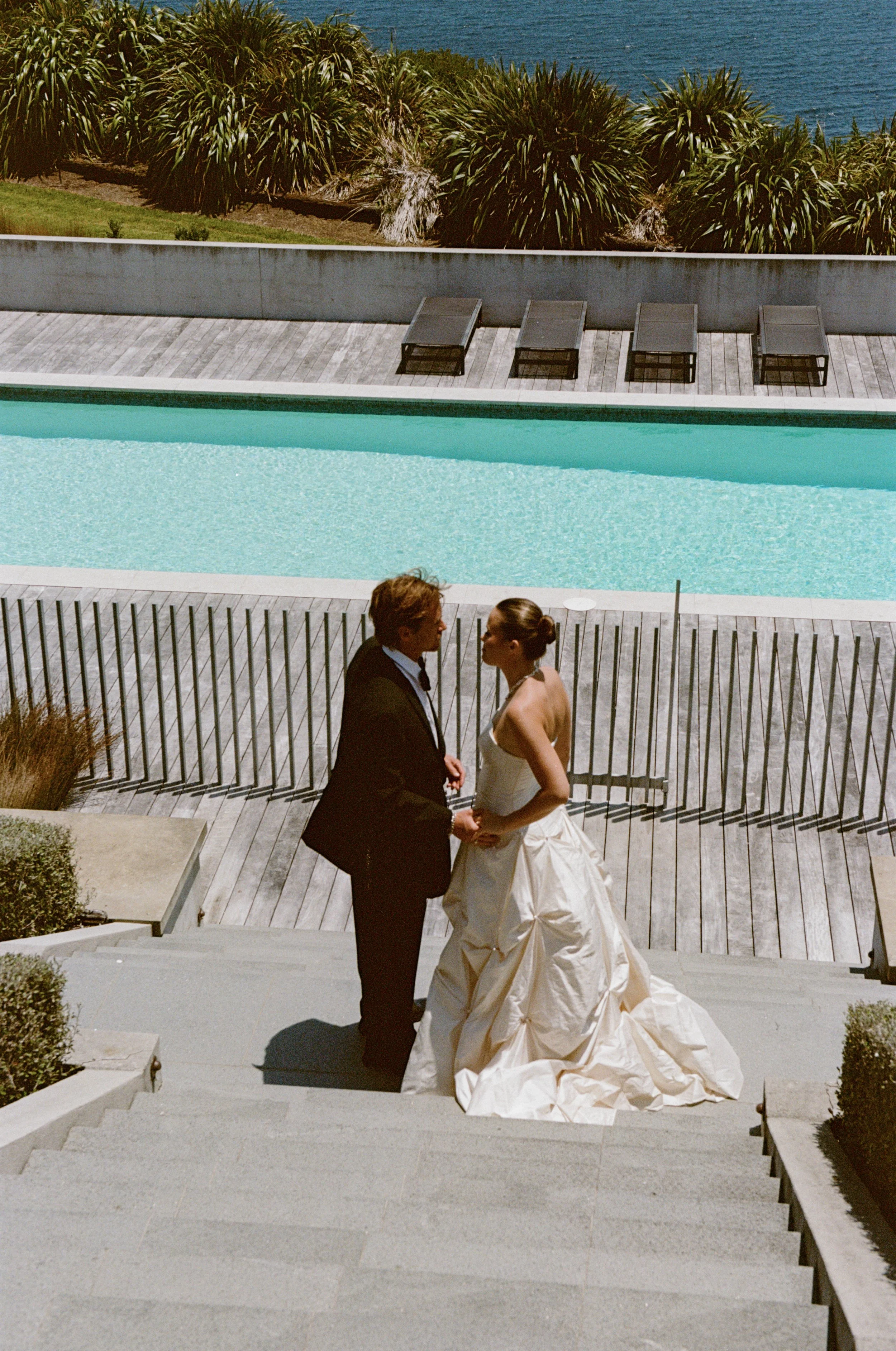 A bride and groom holding hands on a staircase outside near a swimming pool, with lounge chairs and tropical plants in the background.