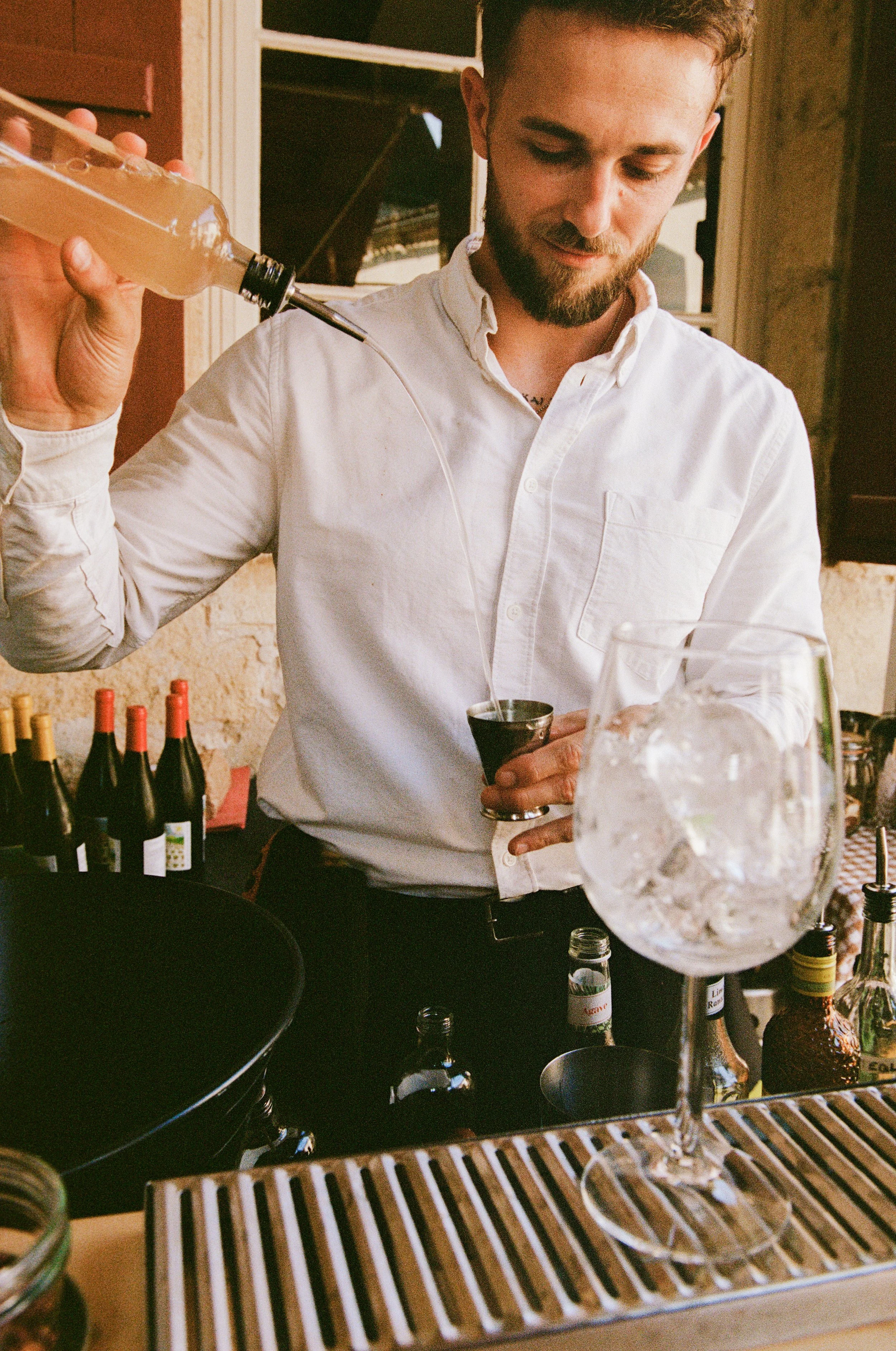 A man in a white shirt pouring a clear liquid from a bottle into a glass at a bar or restaurant setting with wine bottles and glasses on the counter.