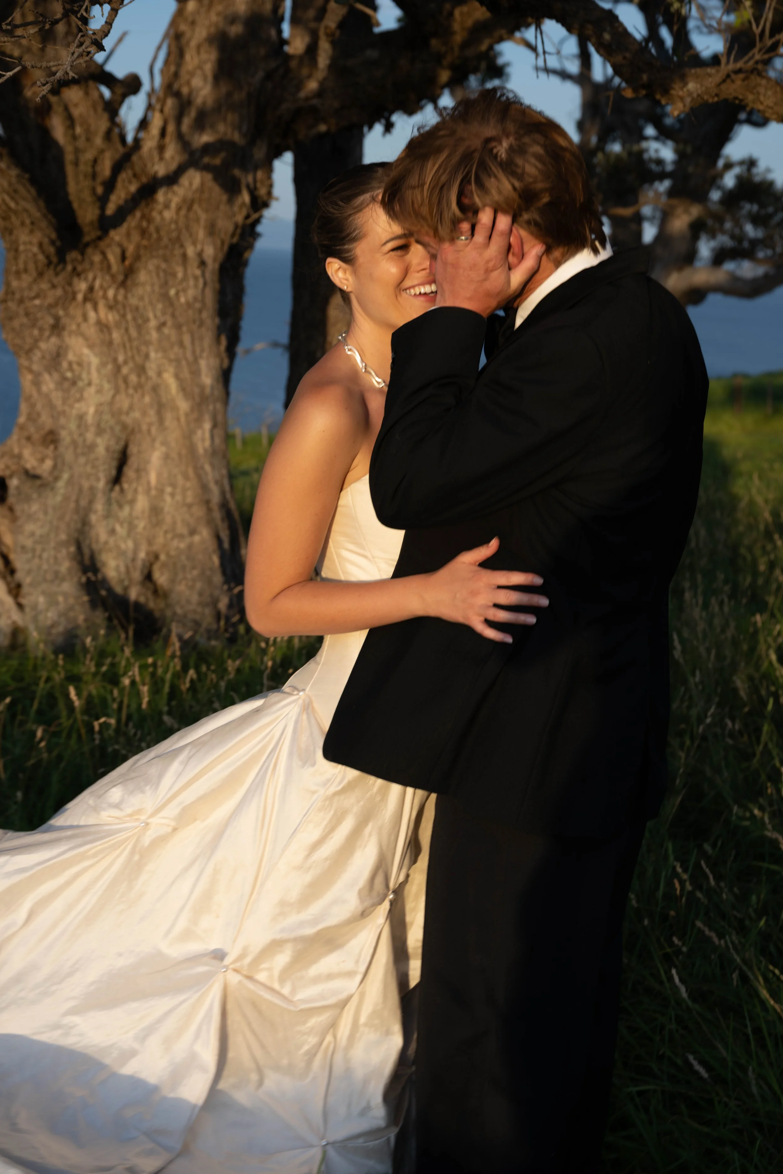 A bride and groom share a kiss outdoors under a tree during their wedding, with the bride in a white gown and the groom in a black suit.