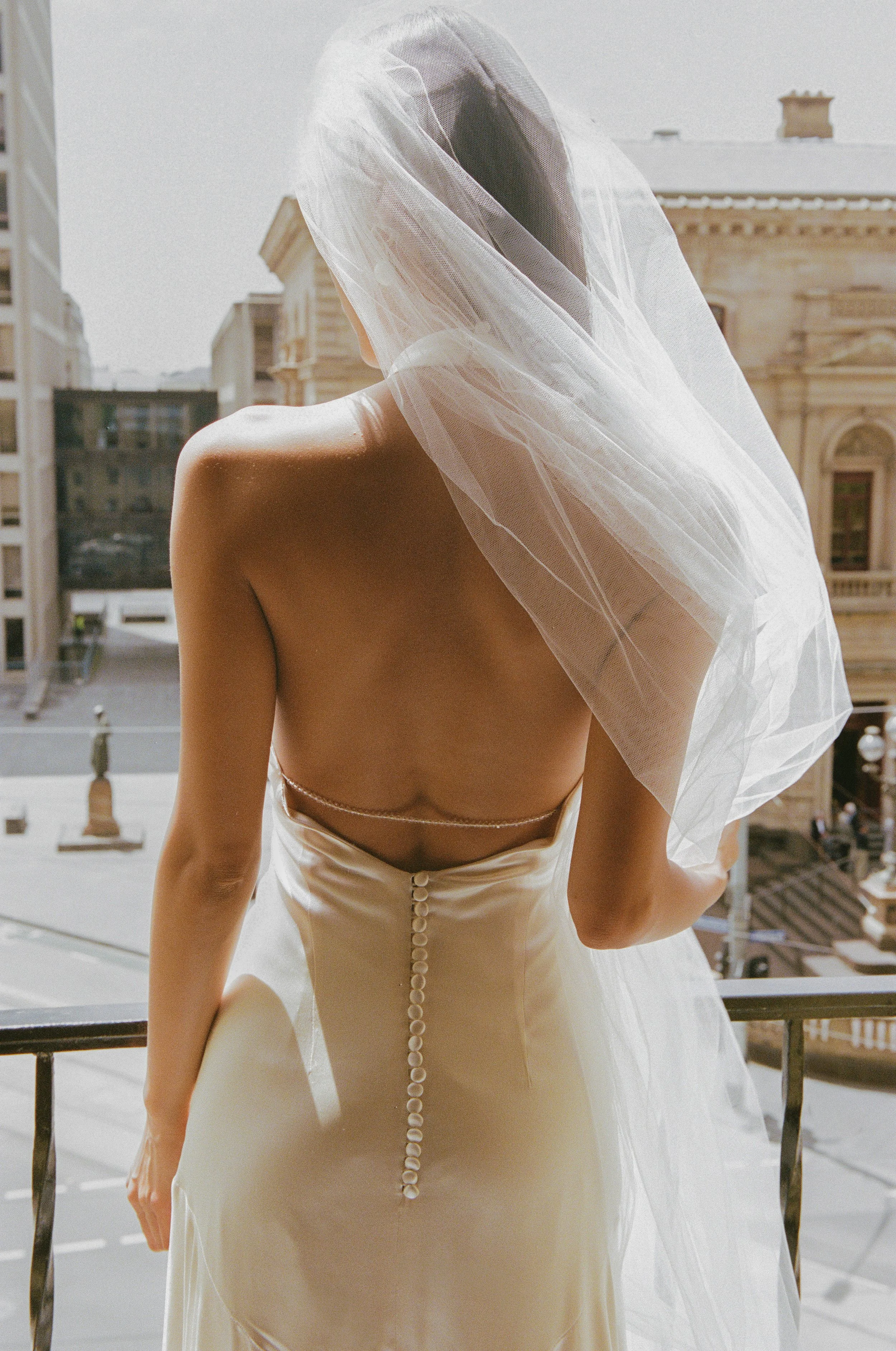 A woman in a wedding dress with her back turned, standing on a balcony with city buildings in the background.