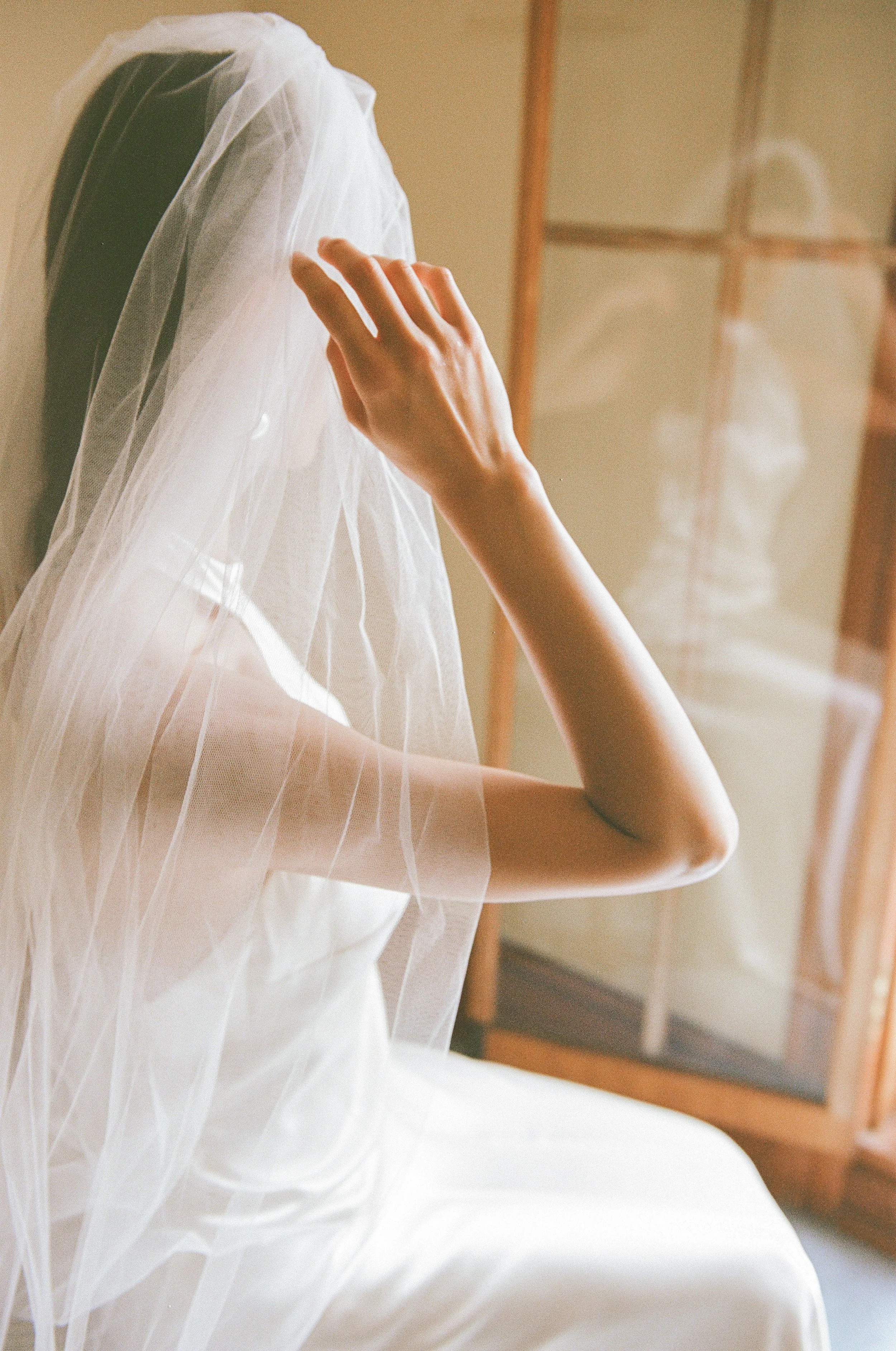A bride wearing a white wedding dress and veil, seen from the side, with her hand touching the veil, sitting in a room with wooden furniture.