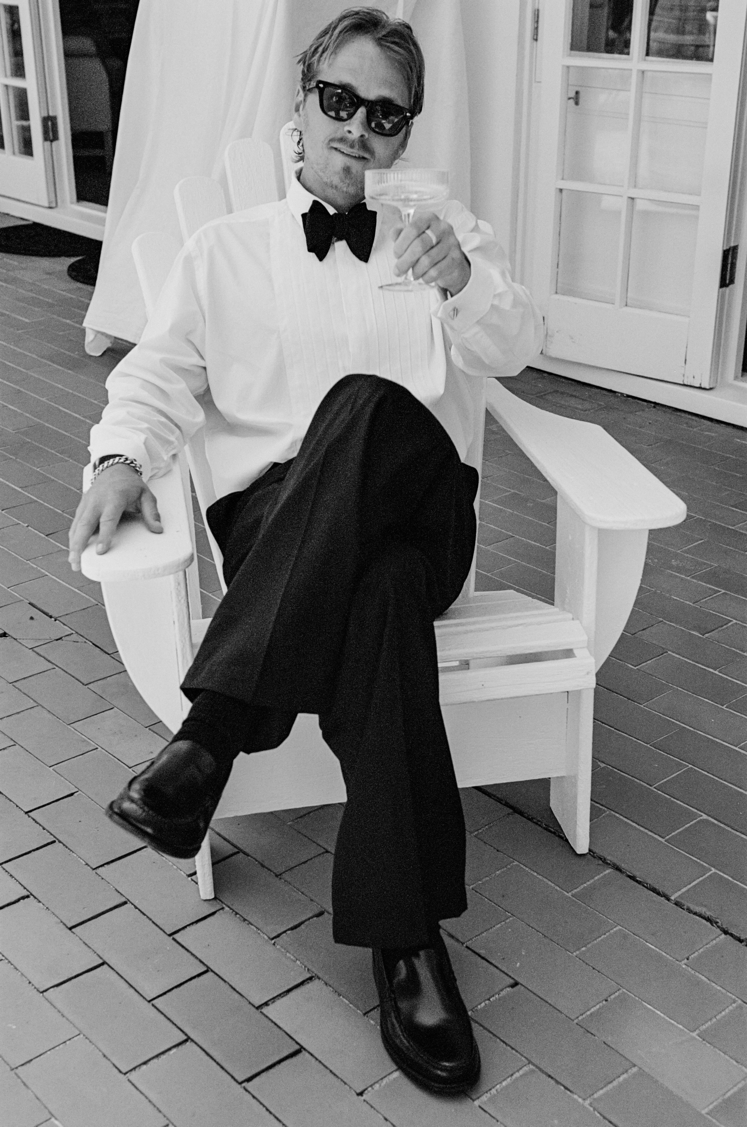 A man wearing sunglasses, a white dress shirt, black bow tie, black pants, and shiny black shoes sitting on a white Adirondack chair, holding a glass in his right hand, with one leg crossed over the other, outdoors on a brick patio near a door with glass panes.