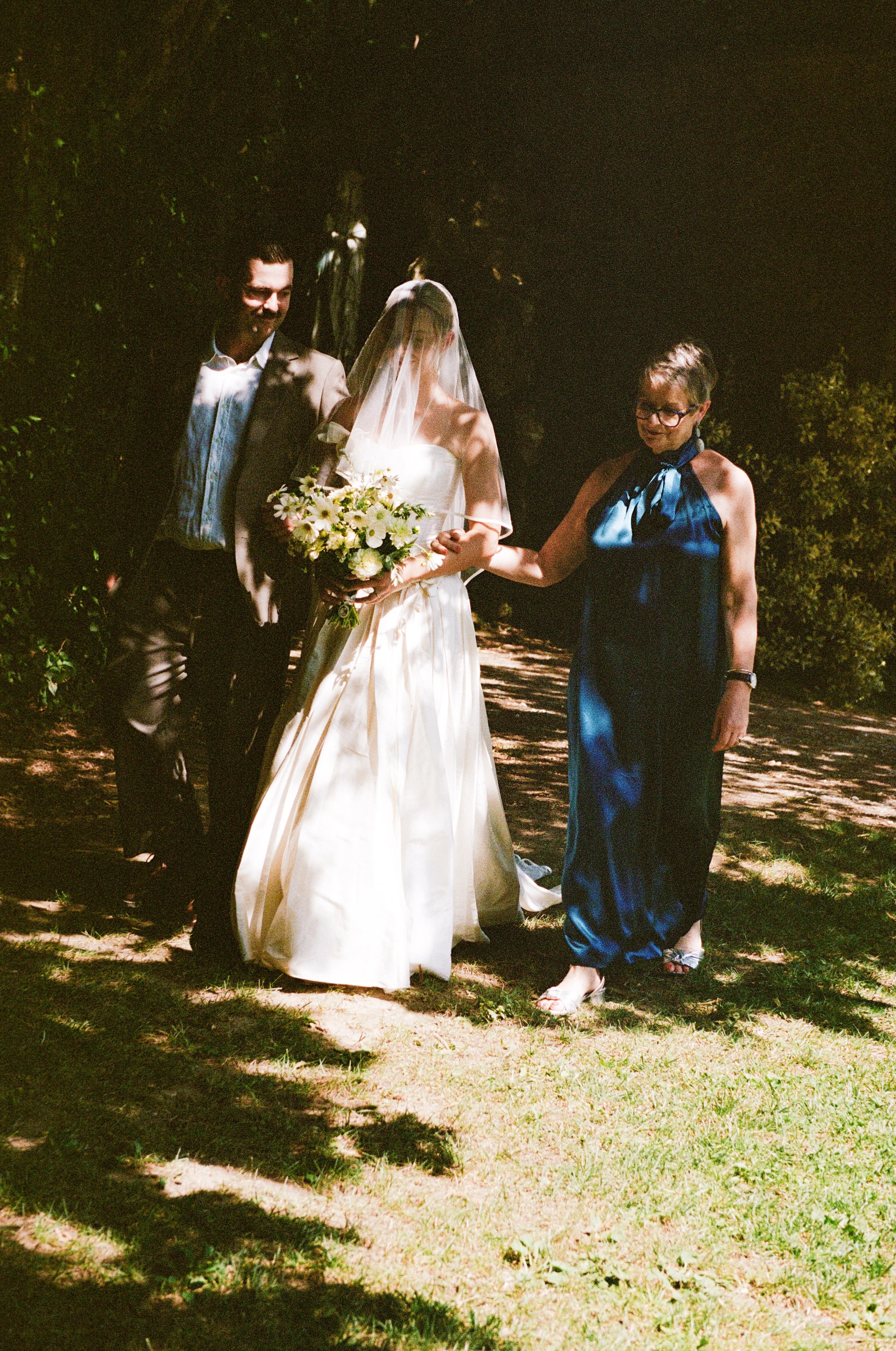 A bride holding a bouquet of white flowers is standing between a man in a beige suit and a woman in a blue dress outdoors at night, with sunlight filtering through trees.