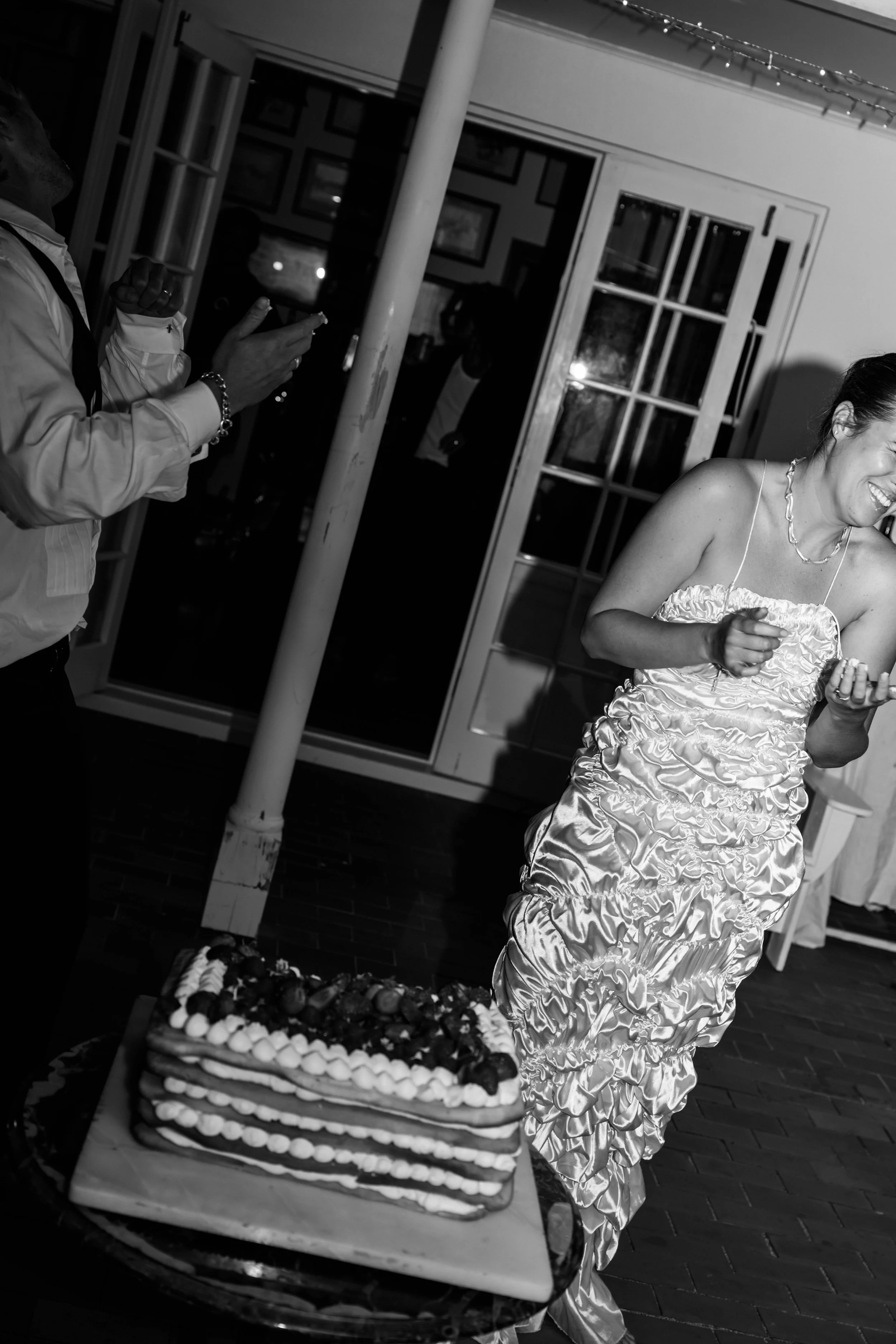 A woman in a ruffled satin dress laughing at a birthday celebration with a layered cake in front of her.
