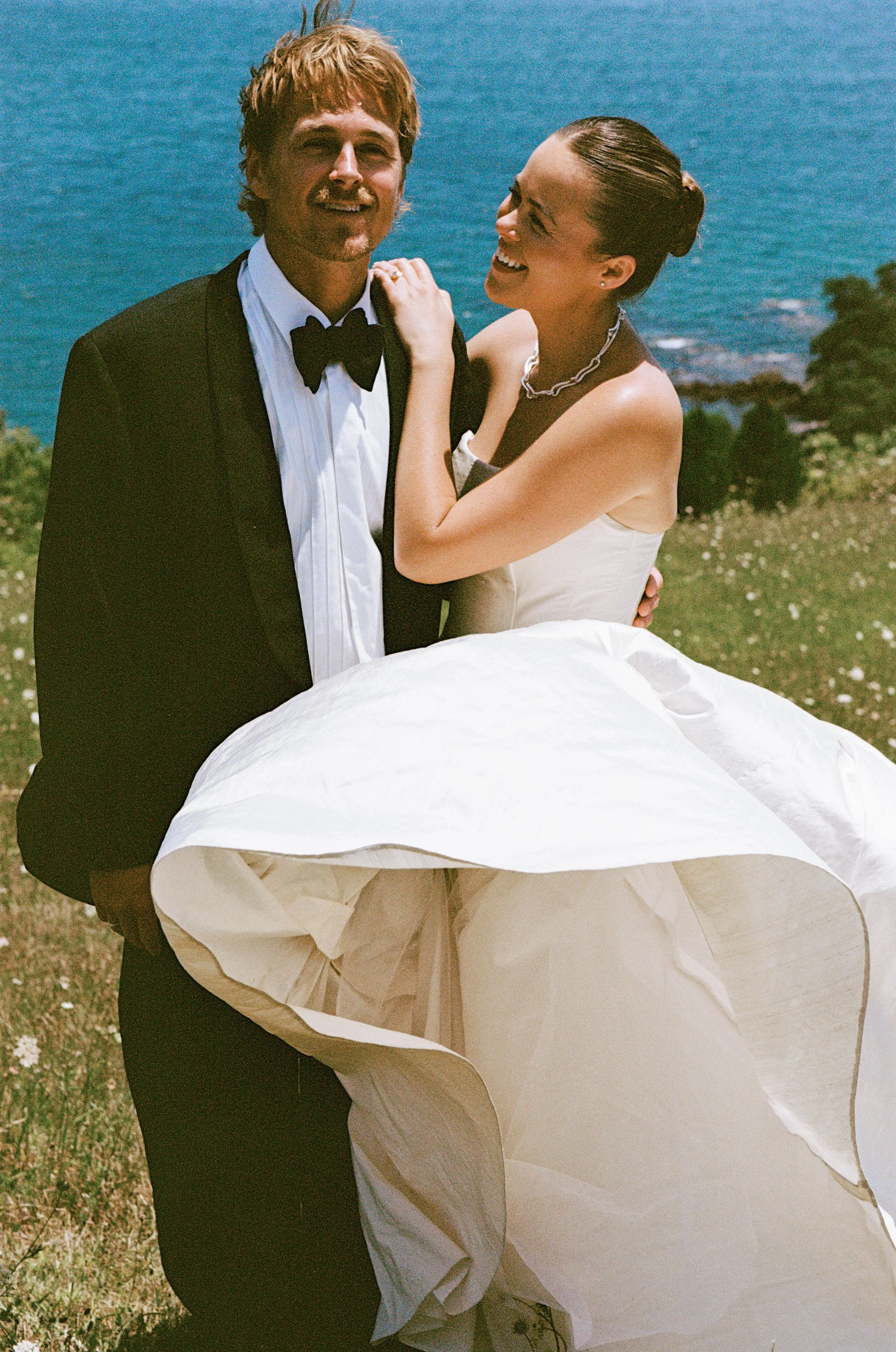 A smiling couple in wedding attire standing outdoors near a body of water, with the woman wearing a strapless white wedding dress and the man in a black tuxedo with a bow tie.