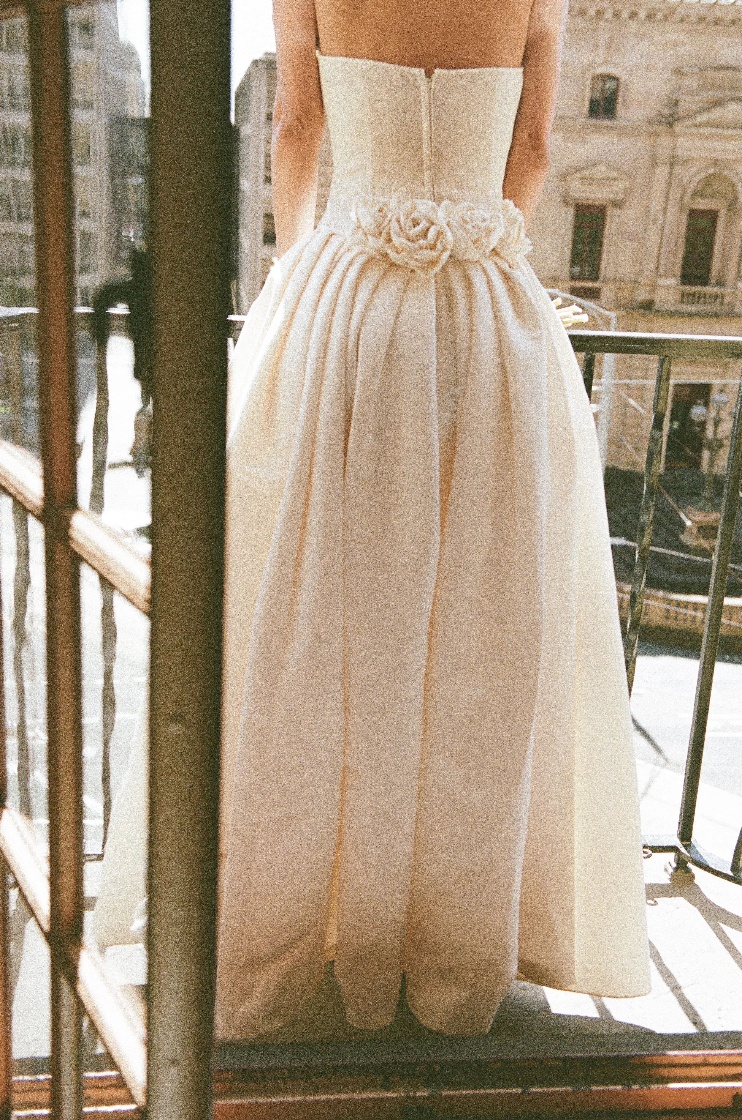 A woman wearing an elegant, strapless white wedding gown with floral embellishments at the waist, standing on a balcony with city buildings in the background.