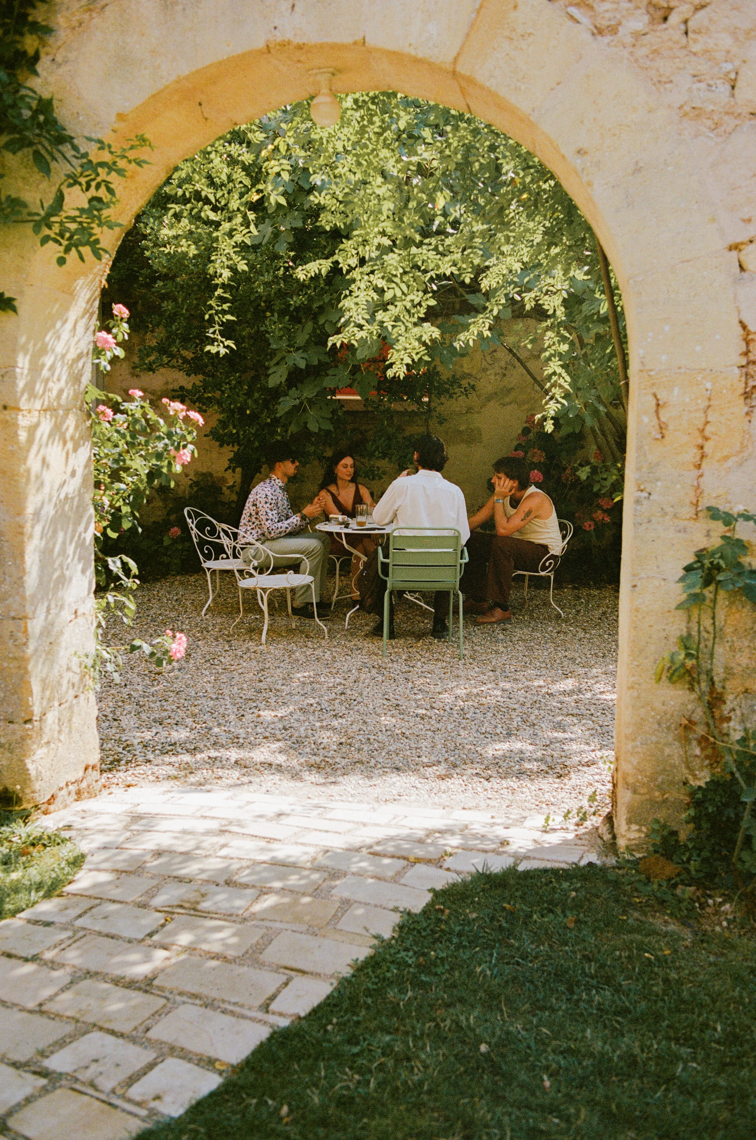 People sitting around a table in a garden courtyard, framed by a stone archway, with trees and flowers in the background.