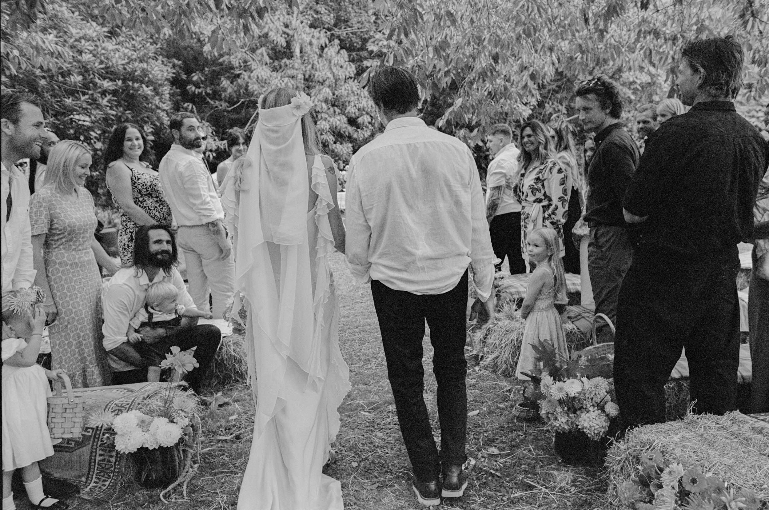 A couple is holding hands and walking down an outdoor aisle during a wedding ceremony, surrounded by family and friends.