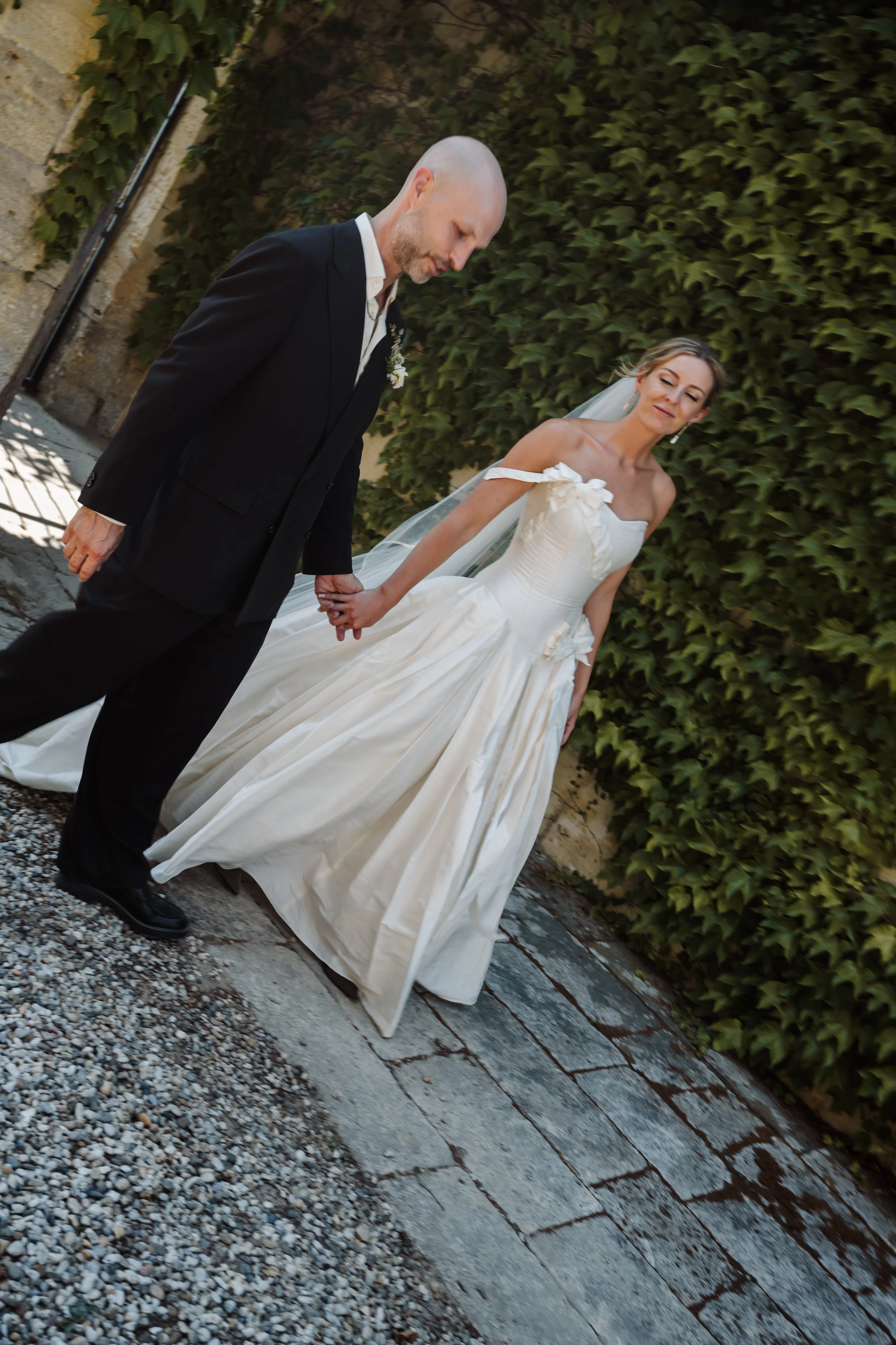 A bride and groom holding hands outdoors, with the groom dressed in a black suit and the bride in a white off-shoulder wedding gown, standing on a path with gravel and stone paving, against a background of green leafy hedge.