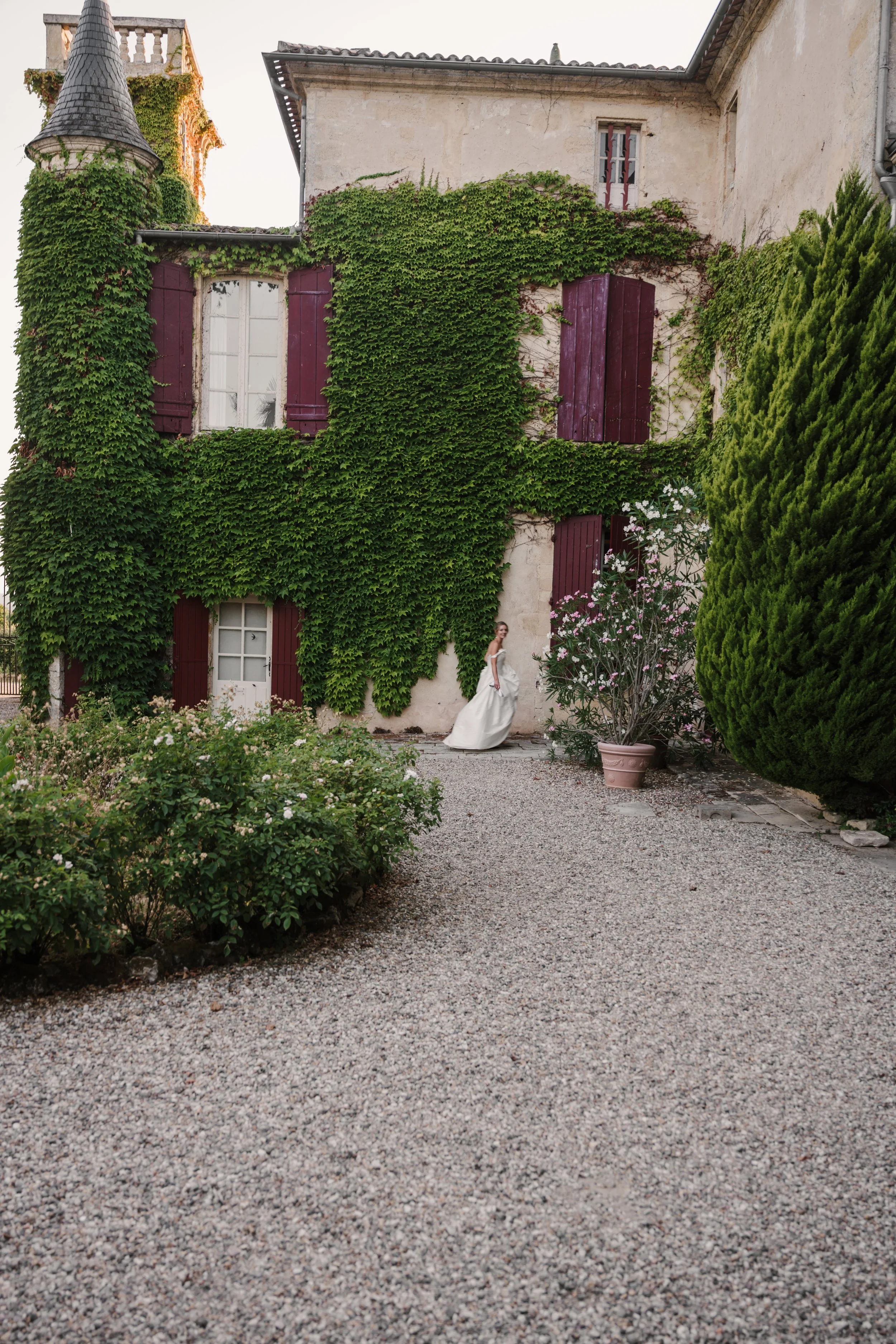 A woman in a white dress standing on a gravel pathway in front of a vine-covered house with red shutters and a small tower with ivy. There are green bushes and flowering plants on the scene.