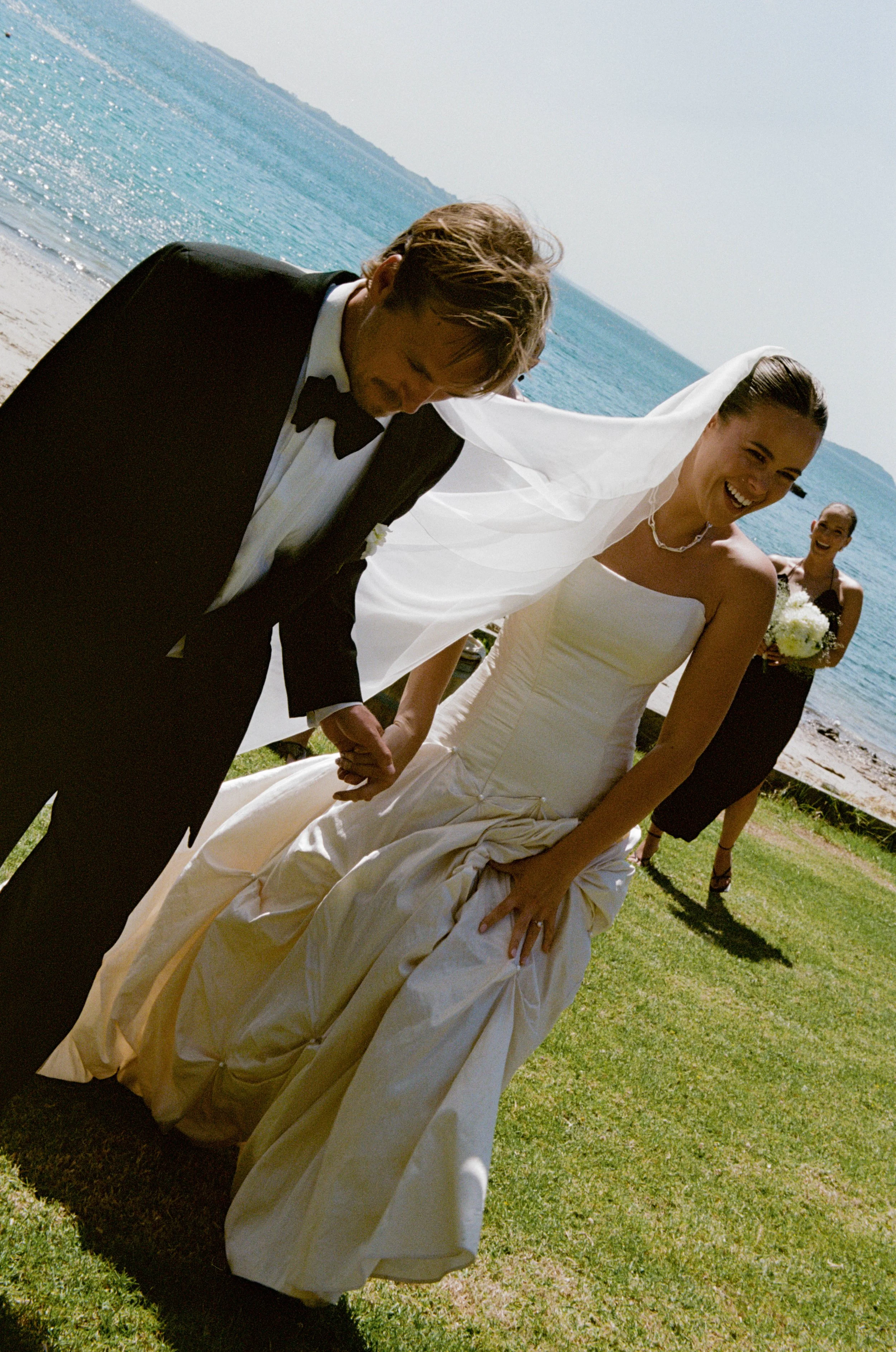 A newly married couple holding hands and smiling on a grassy area by the water, with a bridesmaid holding a bouquet in the background, during a sunny outdoor wedding.