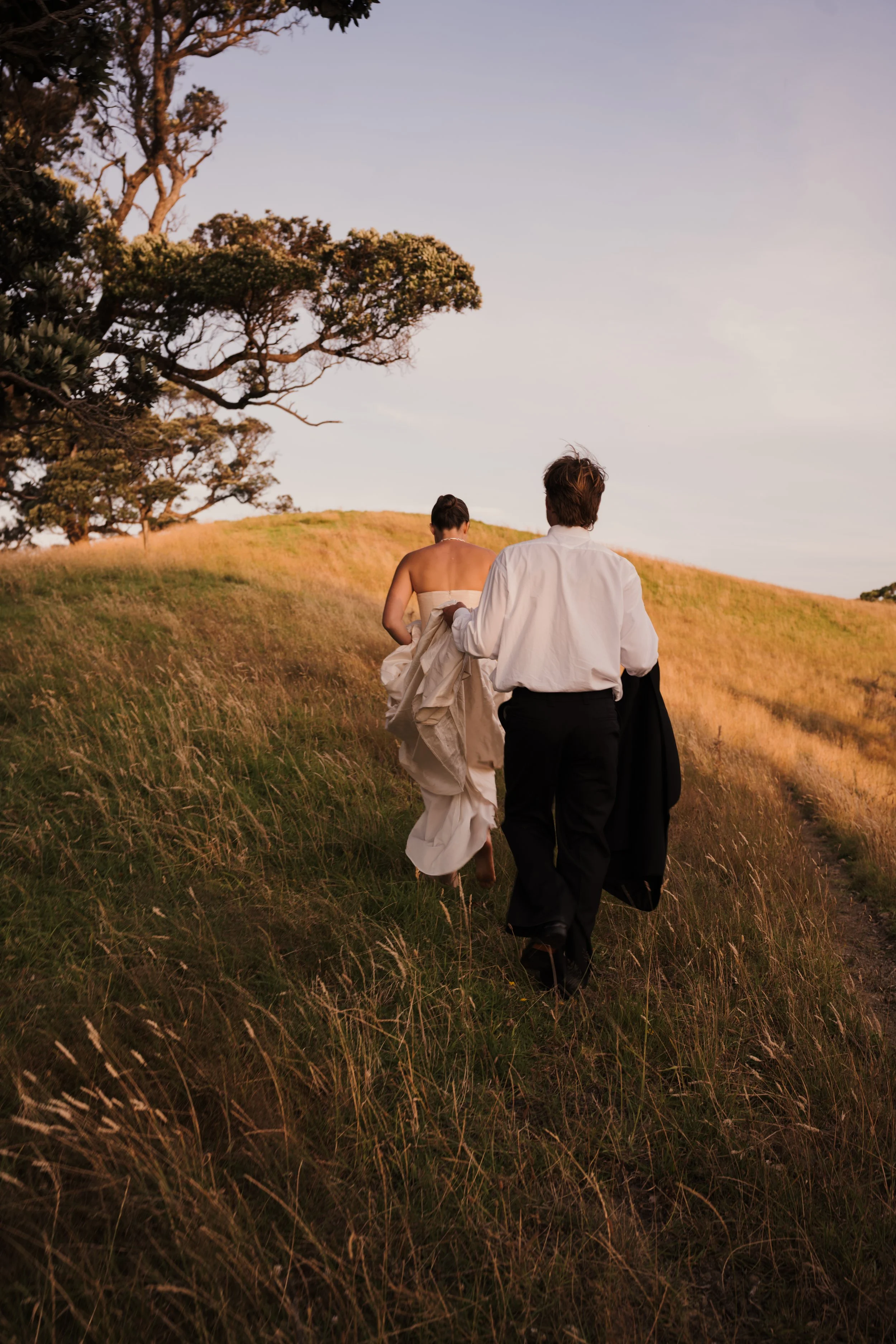 A couple walking uphill through grassy fields at sunset, with the woman in a white wedding dress and the man in a white shirt and dark pants.