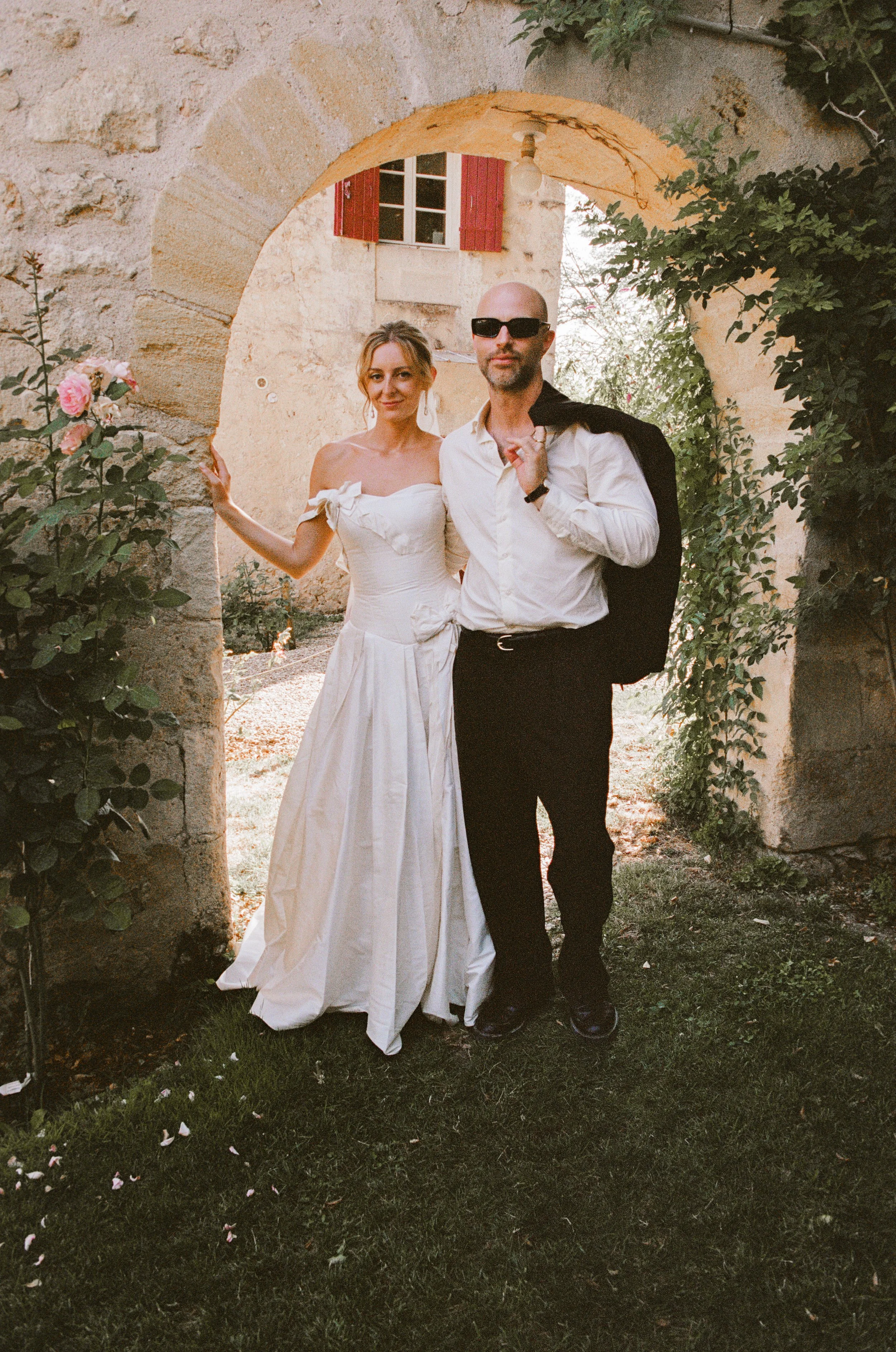 A bride and groom sitting on a log in a grassy field by a large tree, holding beers and taking a photo together.