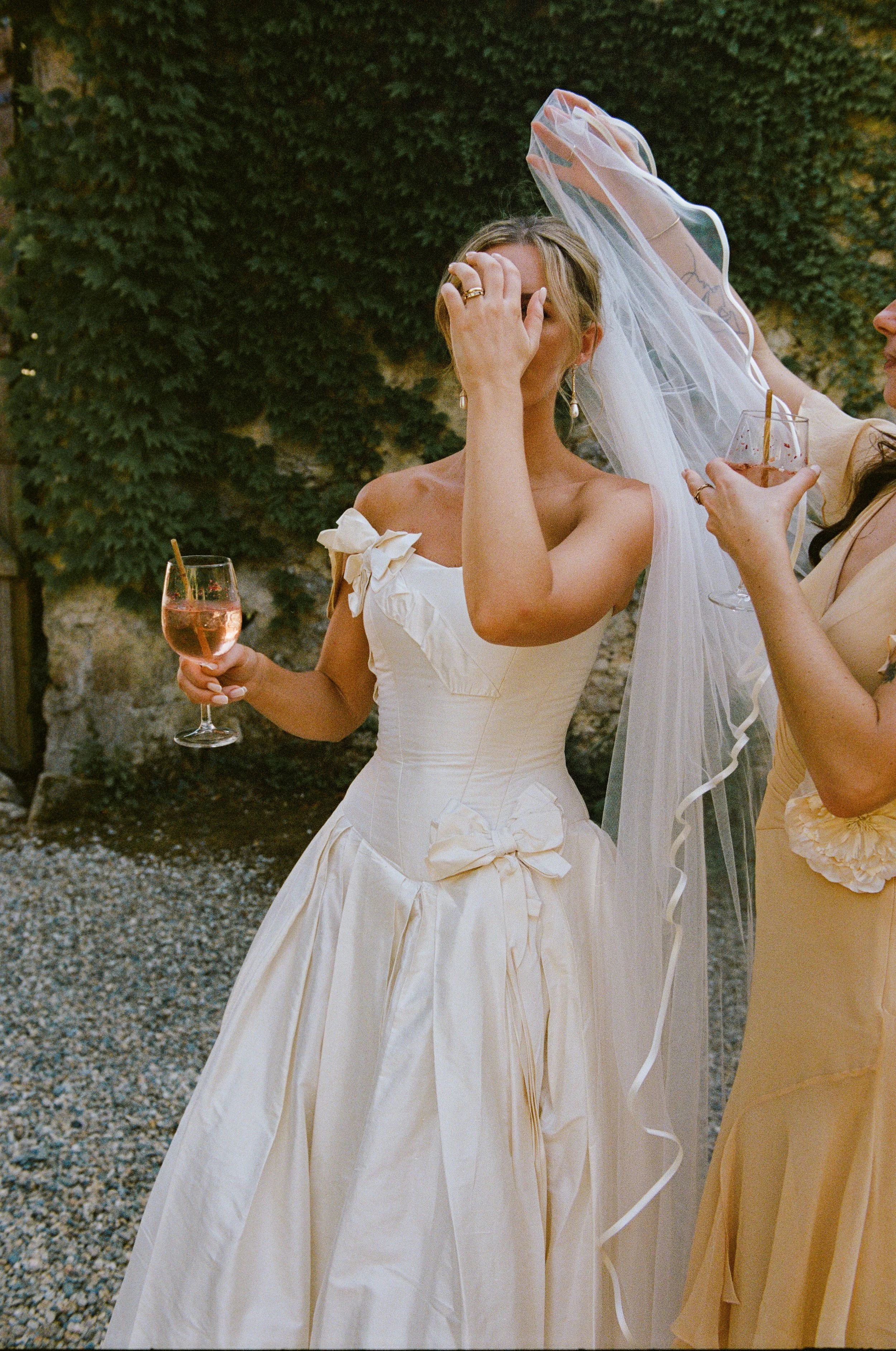 A woman in a wedding dress with a bow on the bodice, holding a glass of pink drink, standing outdoors near a stone wall covered in greenery. Another woman adjusting her veil.