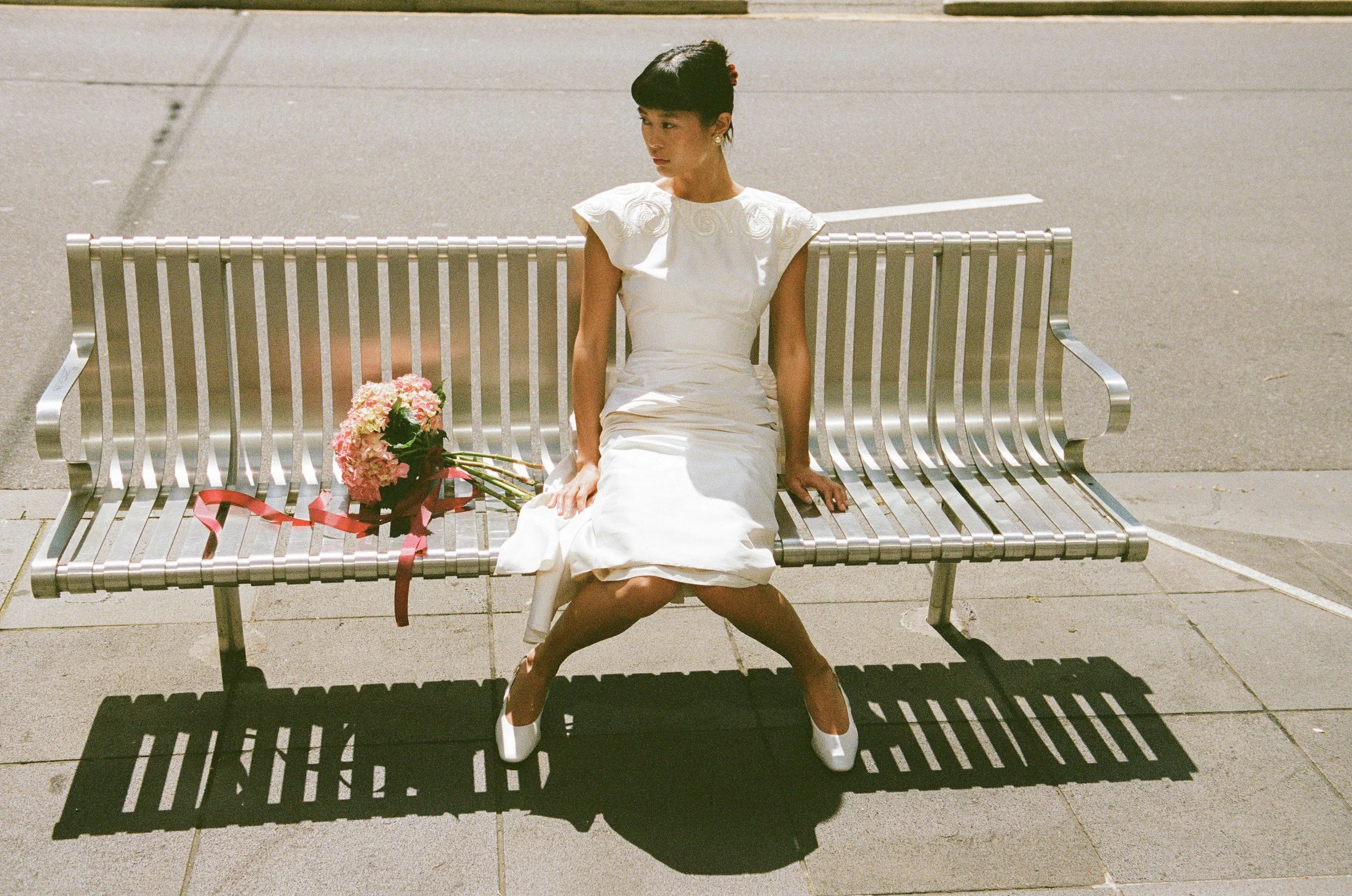 A woman in a white dress sitting on a metal bench with a bouquet of pink flowers next to her, casting a shadow on the sidewalk.