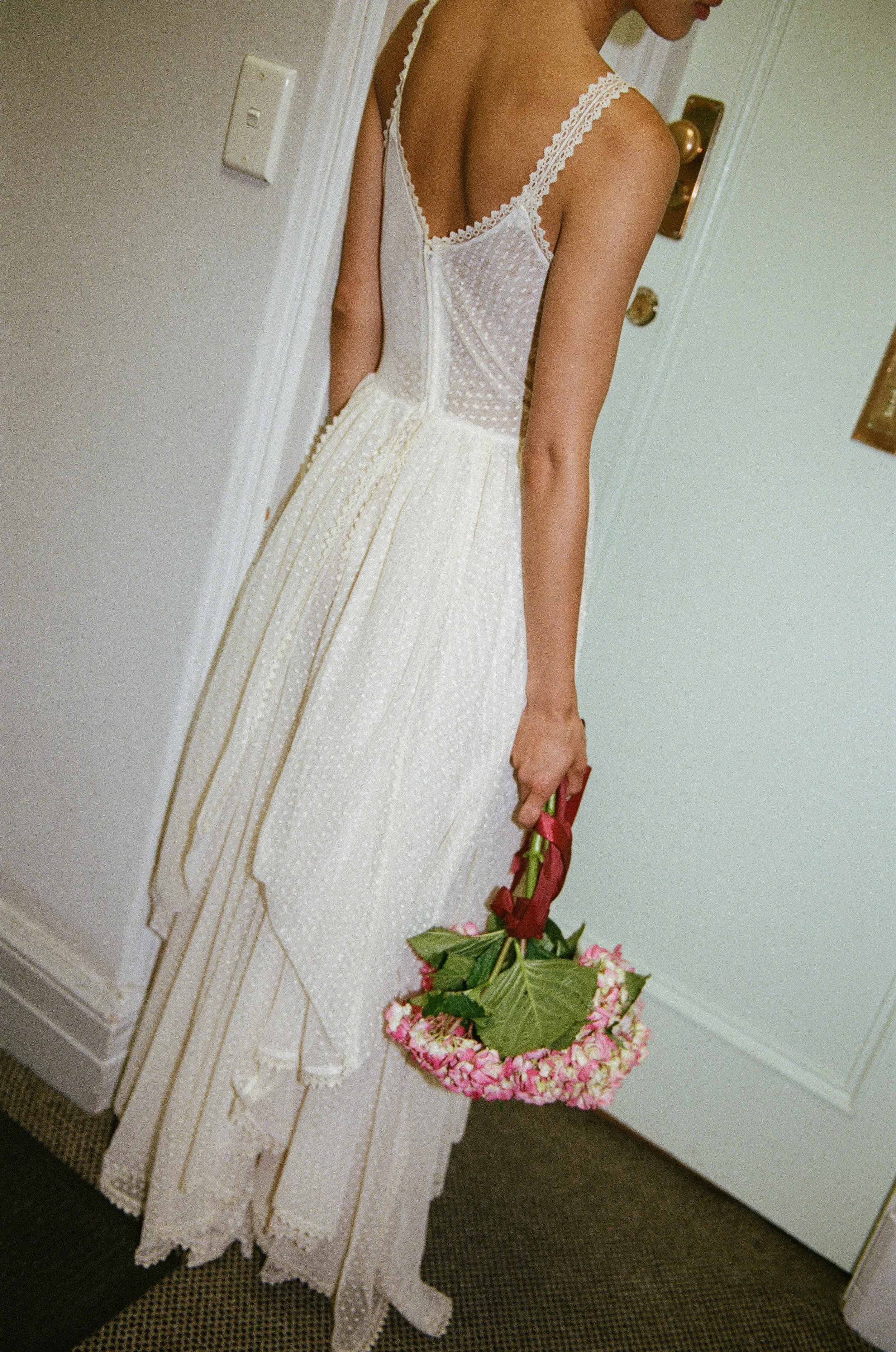 A woman in a vintage white dress holding a pink flower-embellished basket, standing by a door.