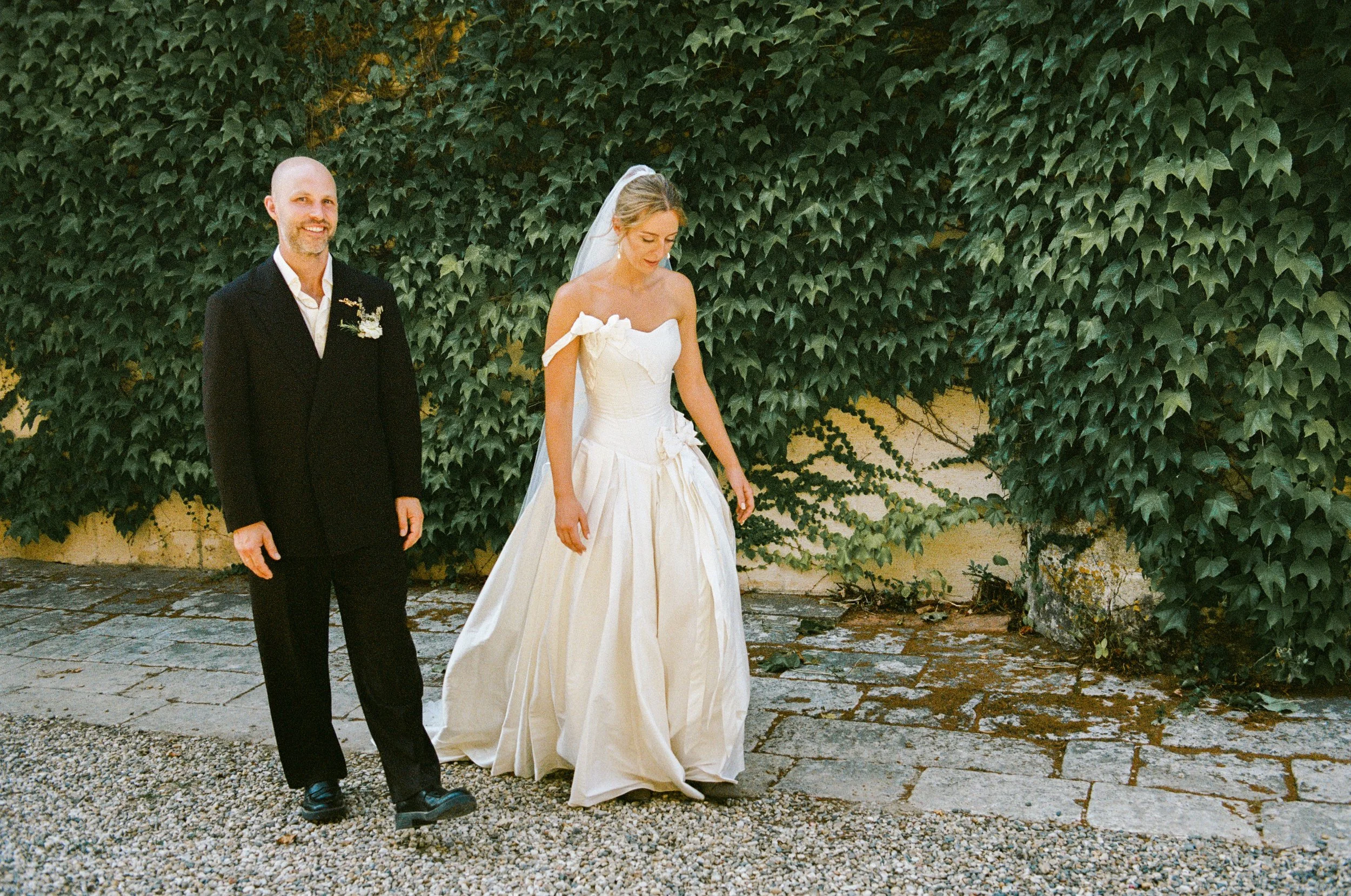 A wedding scene with a groom in a black suit smiling and a bride in a white wedding dress looking down, standing outdoors in front of a green ivy-covered wall.