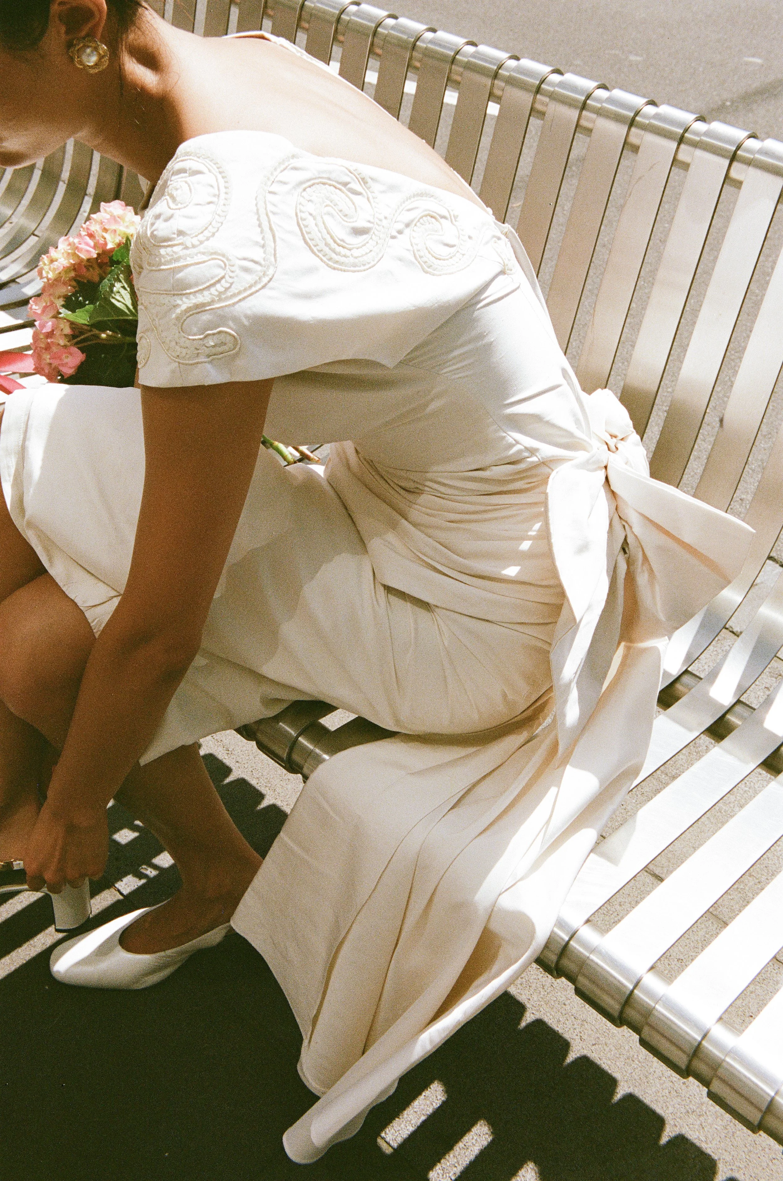 A woman wearing a white dress with embroidery on the shoulder, sitting on a metal bench, adjusting her white high heels, holding a bouquet of pink flowers.