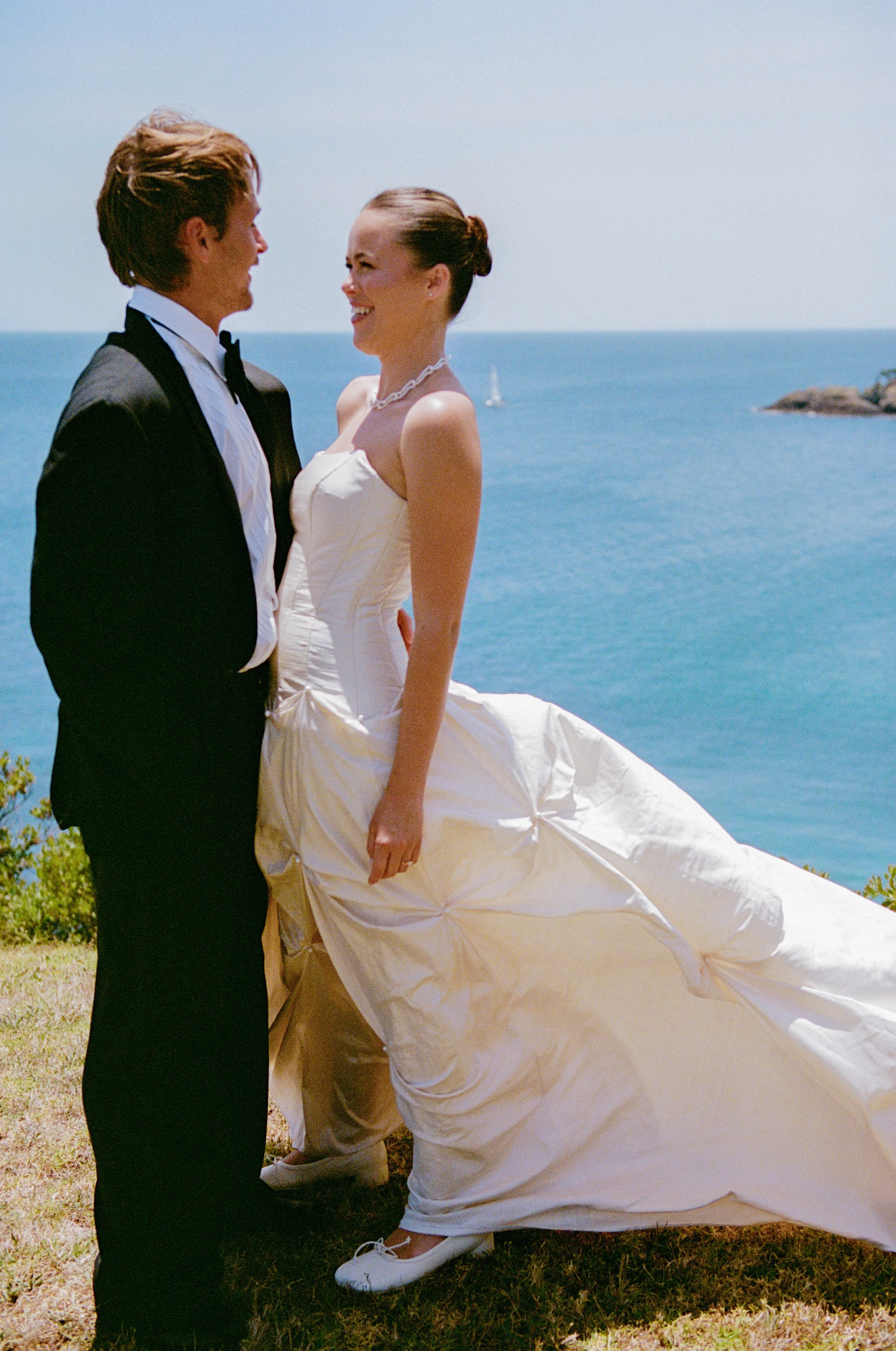 A smiling bride and groom standing outside near the ocean, holding hands and gazing into each other's eyes on their wedding day.