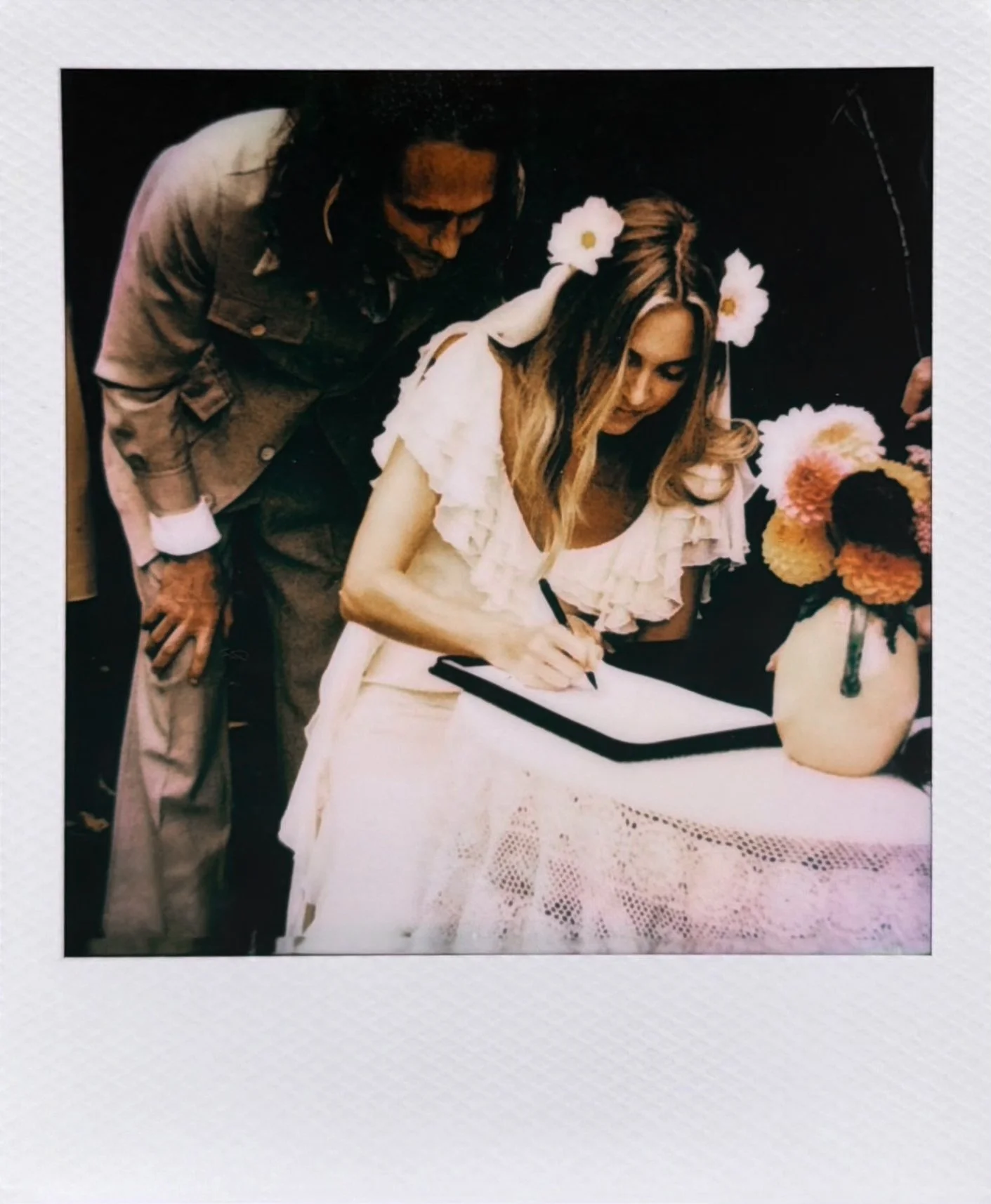 A woman with long hair and flower accessories on her head is signing a document at a table, while a man is leaning over her.