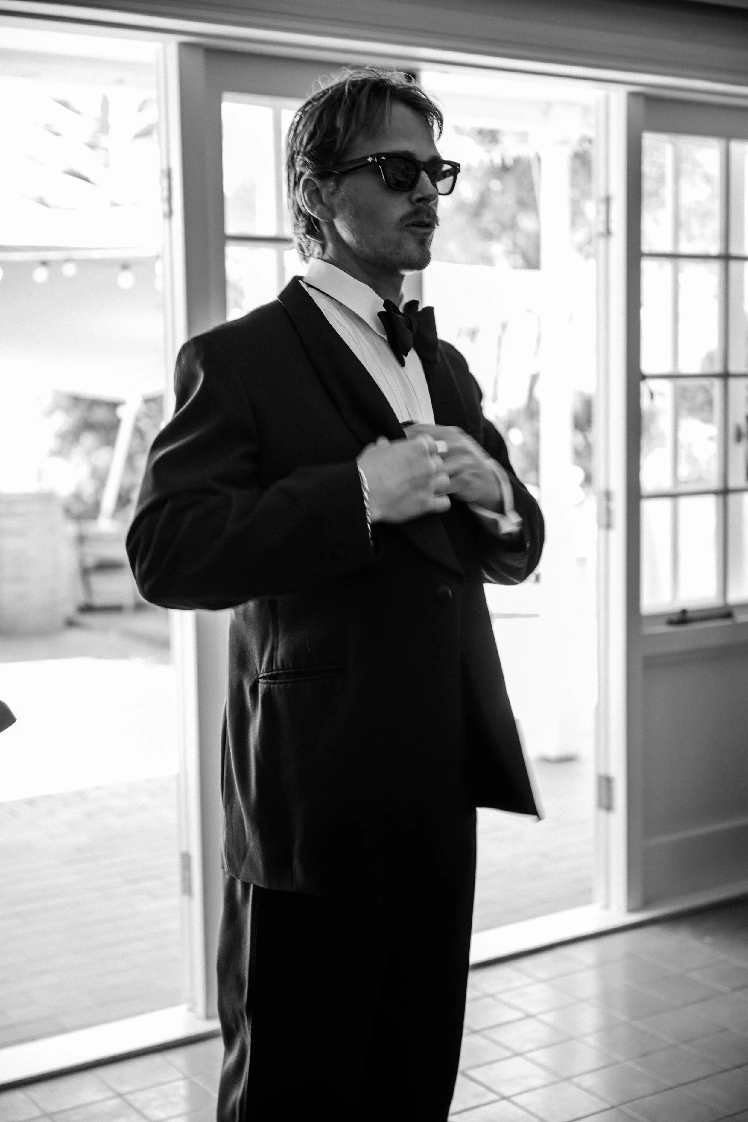 A man in a tuxedo with a bowtie and sunglasses adjusting his jacket indoors with a sliding glass door and patio in the background.