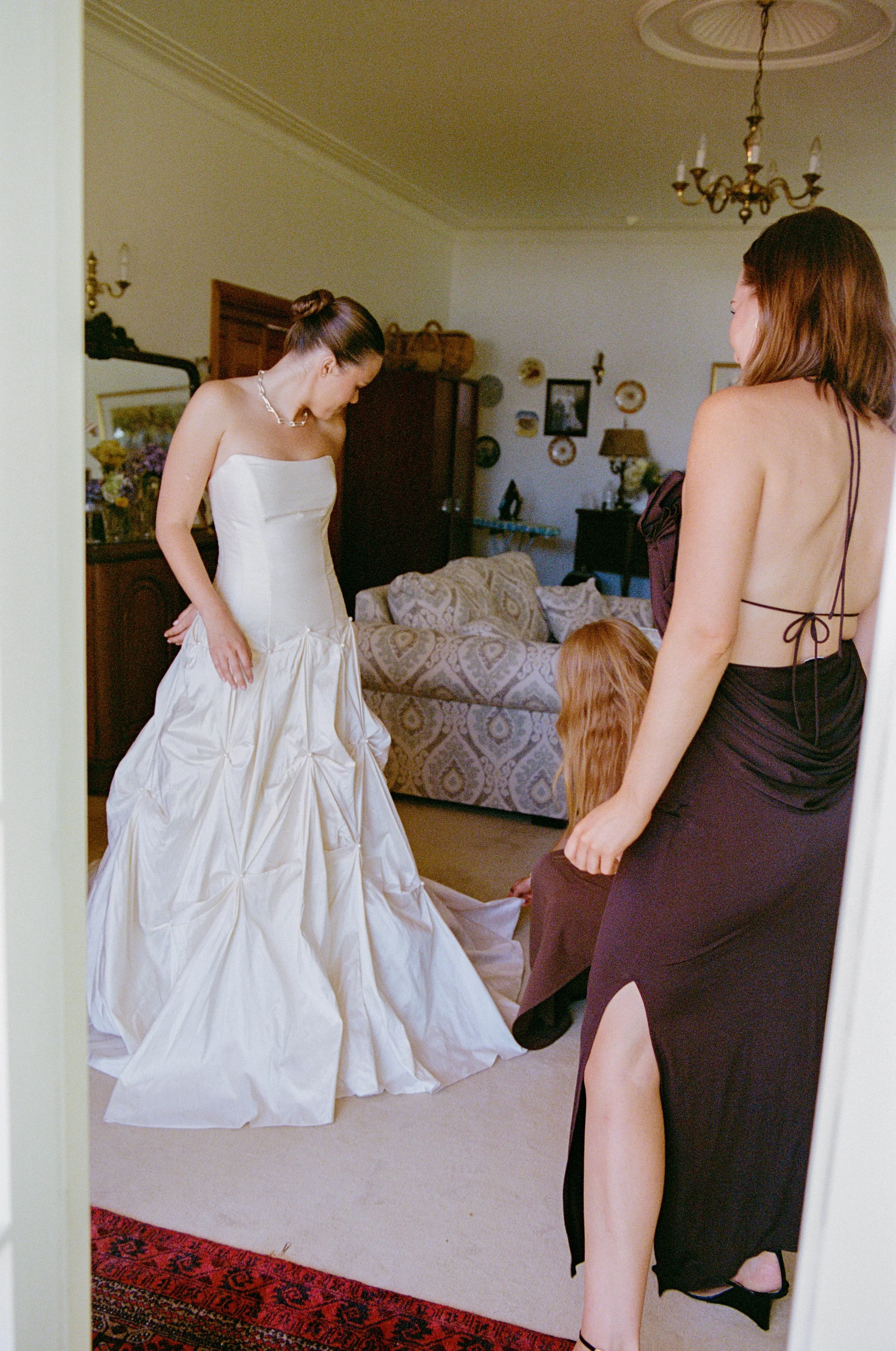 A bride in a strapless wedding gown is standing in a living room, while two women help her with her dress. One woman, in a black dress with a back slit, is standing and another woman with red hair is kneeling on the floor adjusting the gown.