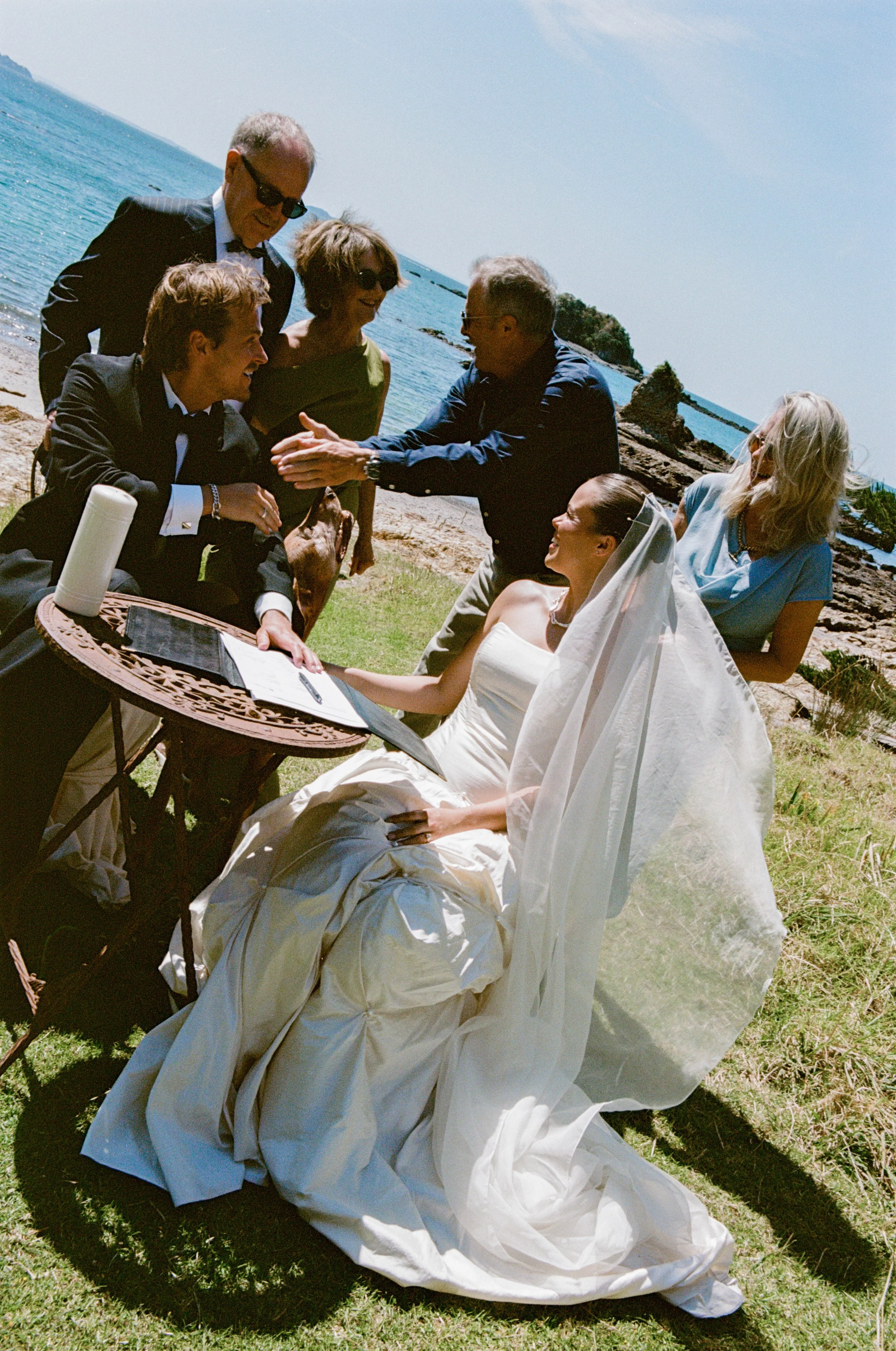 A wedding ceremony taking place outdoors by the ocean. The bride in a white dress and veil is seated, while the groom in a black tuxedo is signing a document at a small table. Guests are gathered around, celebrating and clapping, with a rocky shoreline and blue sea in the background.