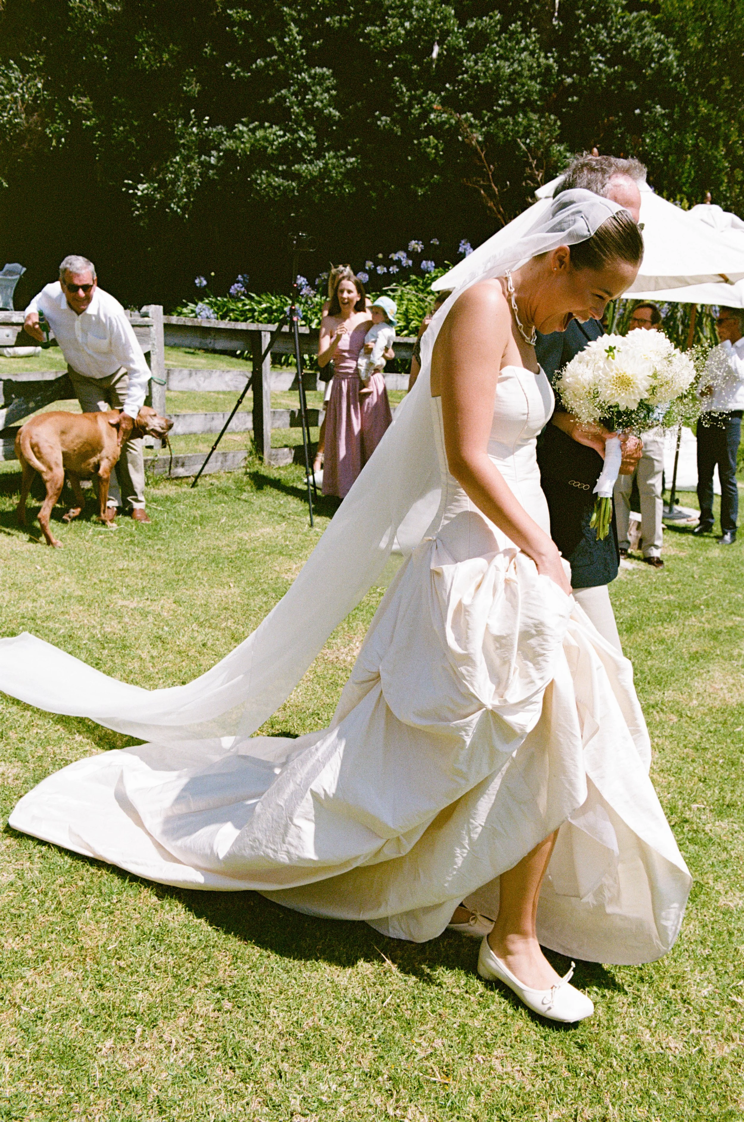 A bride in a white wedding gown and veil holds up her dress as she walks outdoors on a green lawn, smiling. Guests and a dog are visible in the background.