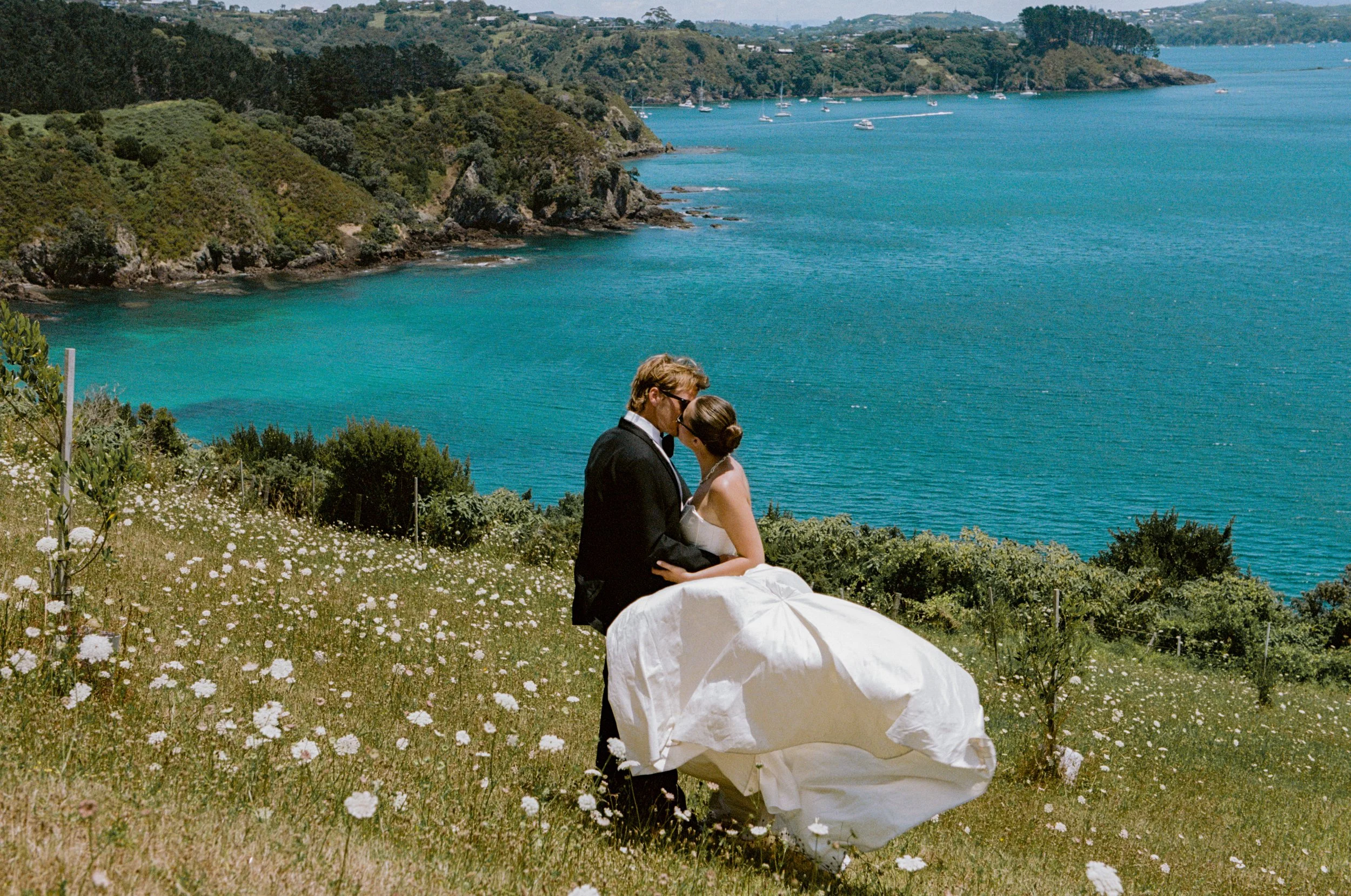 A couple in wedding attire kissing on a grassy hillside with white flowers, overlooking a large blue body of water with boats and green hills in the background.