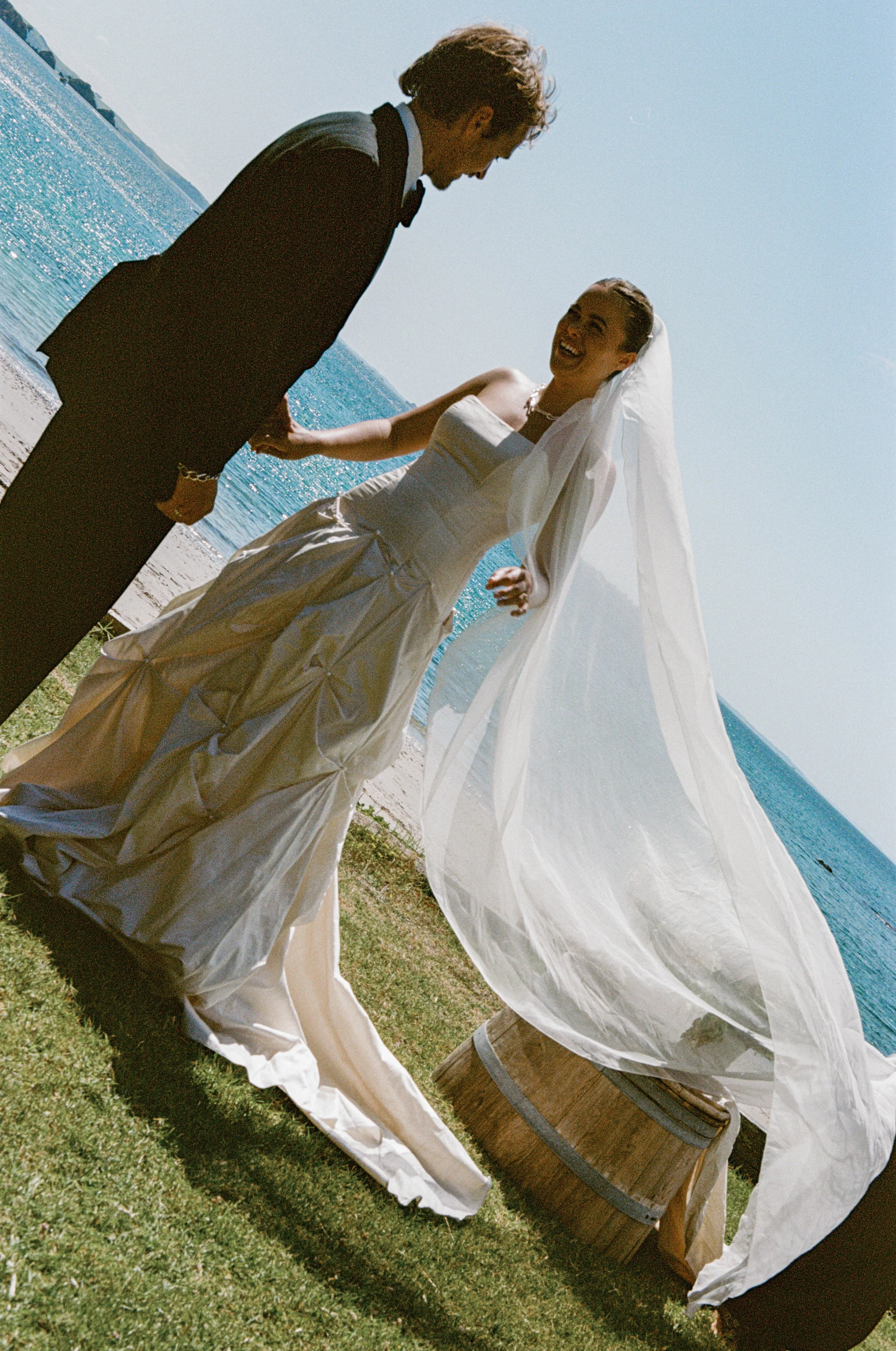 A bride and groom holding hands by the water at a beach, smiling and enjoying their wedding day.