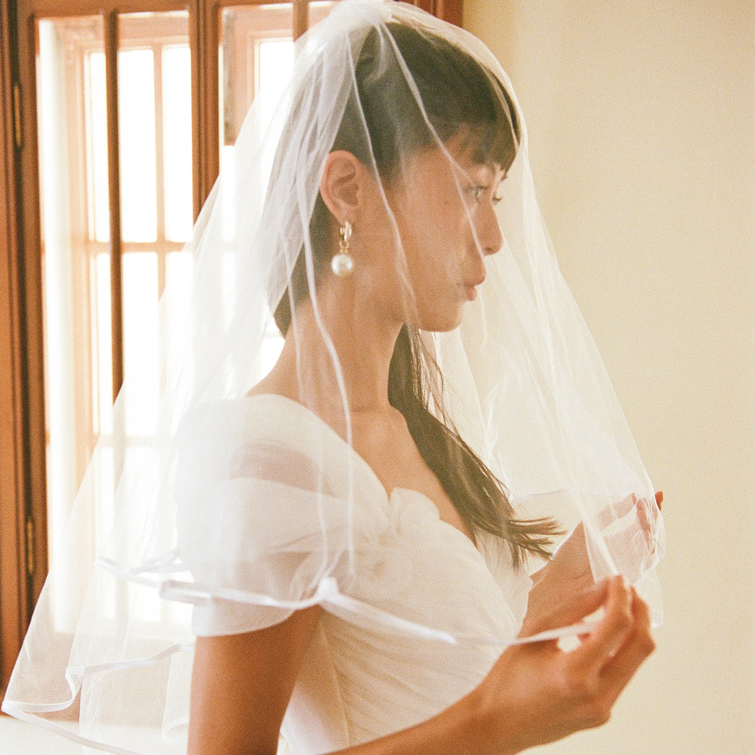 A bride with dark hair wearing a white wedding dress and large pearl earrings, standing indoors near a window with wooden frames, holding a piece of paper or card under a sheer veil.