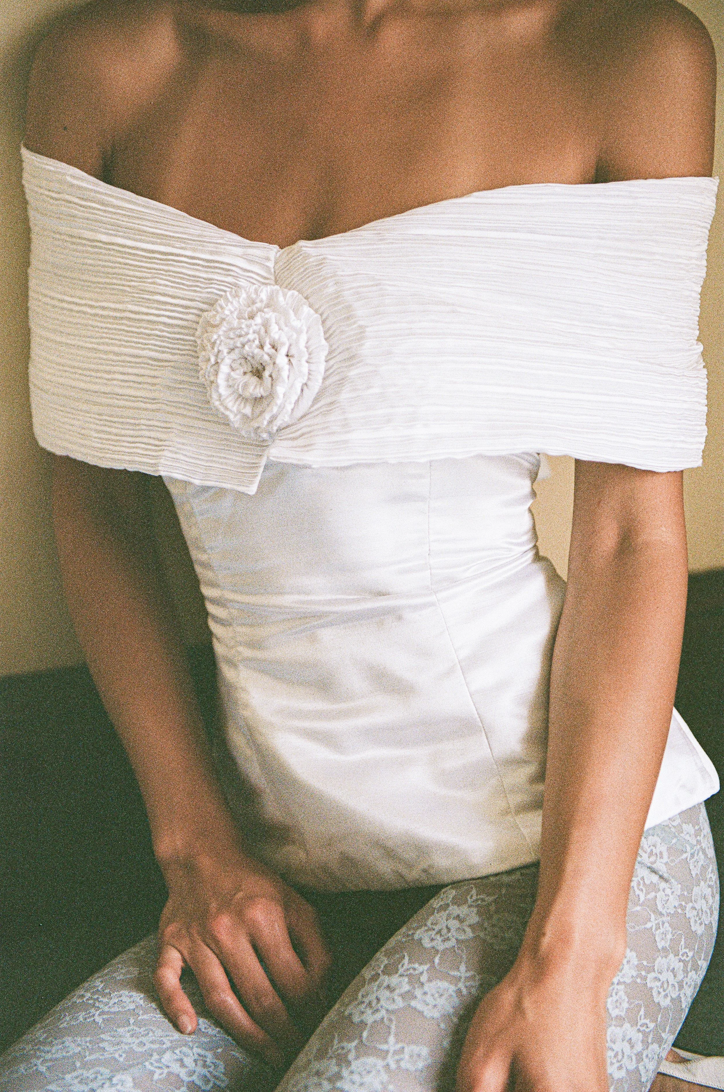A woman wearing an off-shoulder white dress with a large floral embellishment on the front, sitting with her hands on her lap, and wearing lace patterned tights.