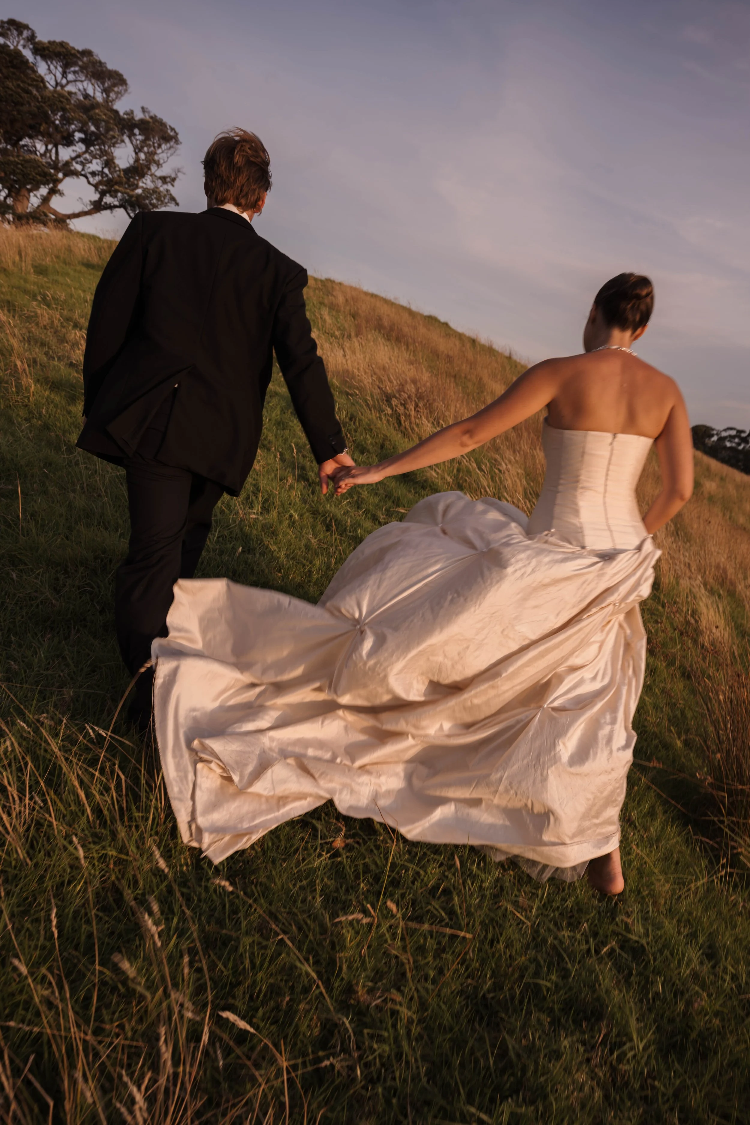 A man and woman holding hands, walking through a grassy field at sunset. The woman is wearing a white wedding dress, and the man is dressed in a black suit.