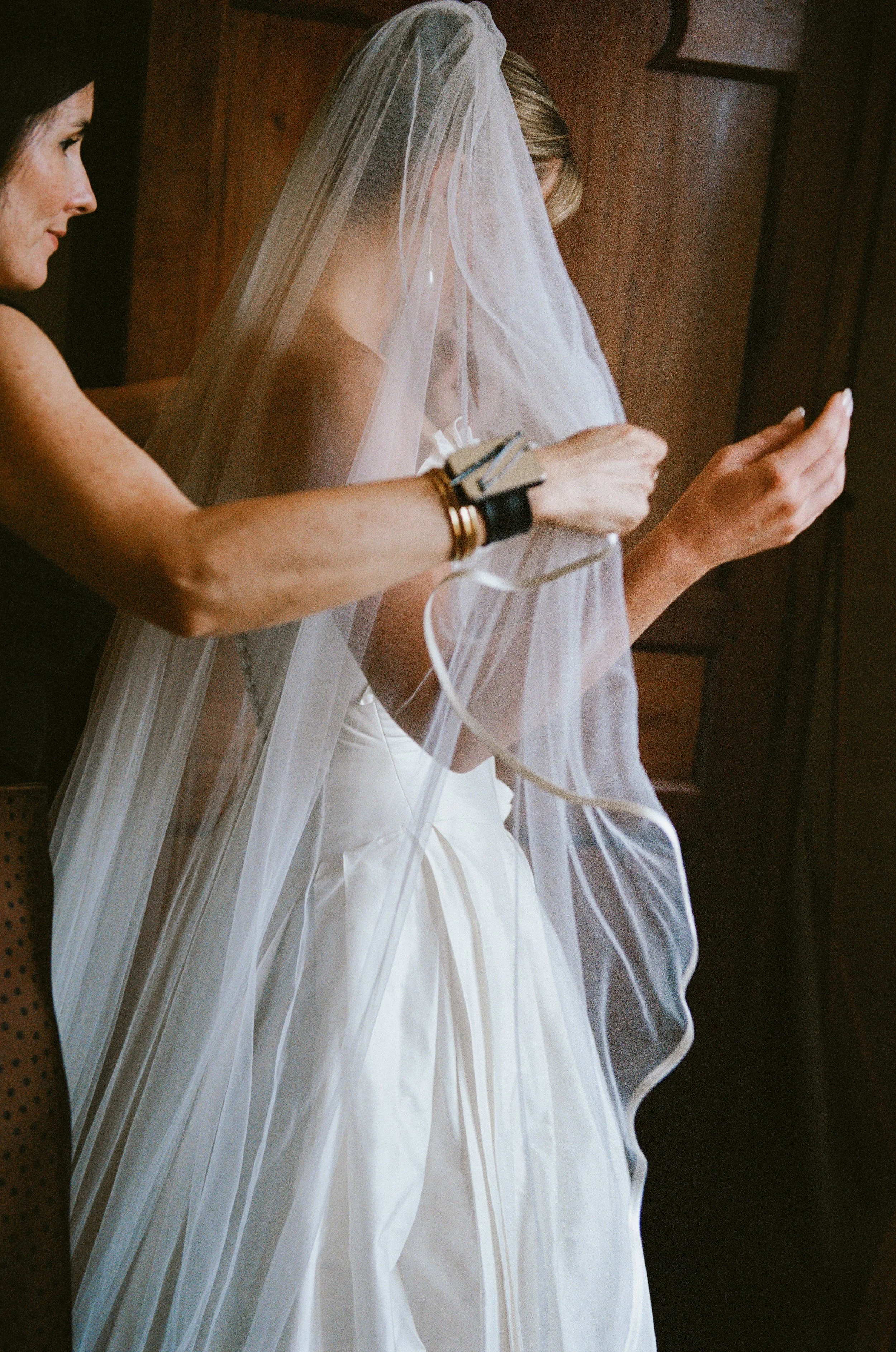A bride in a white wedding dress and veil standing indoors, with a woman assisting her, possibly fitting or adjusting the dress.