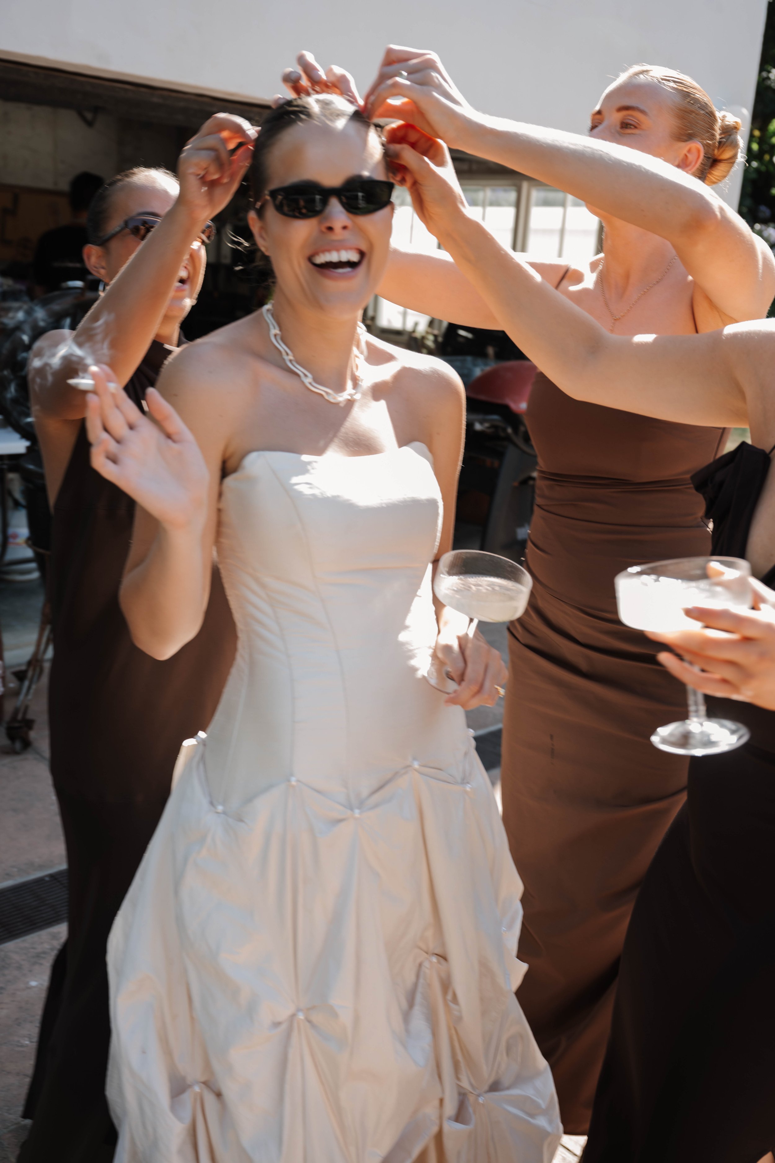A woman in a white wedding dress wearing sunglasses and a pearl necklace is smiling while holding a drink. She is surrounded by women in dresses who are fixing her hair during a celebration outdoors in daylight.