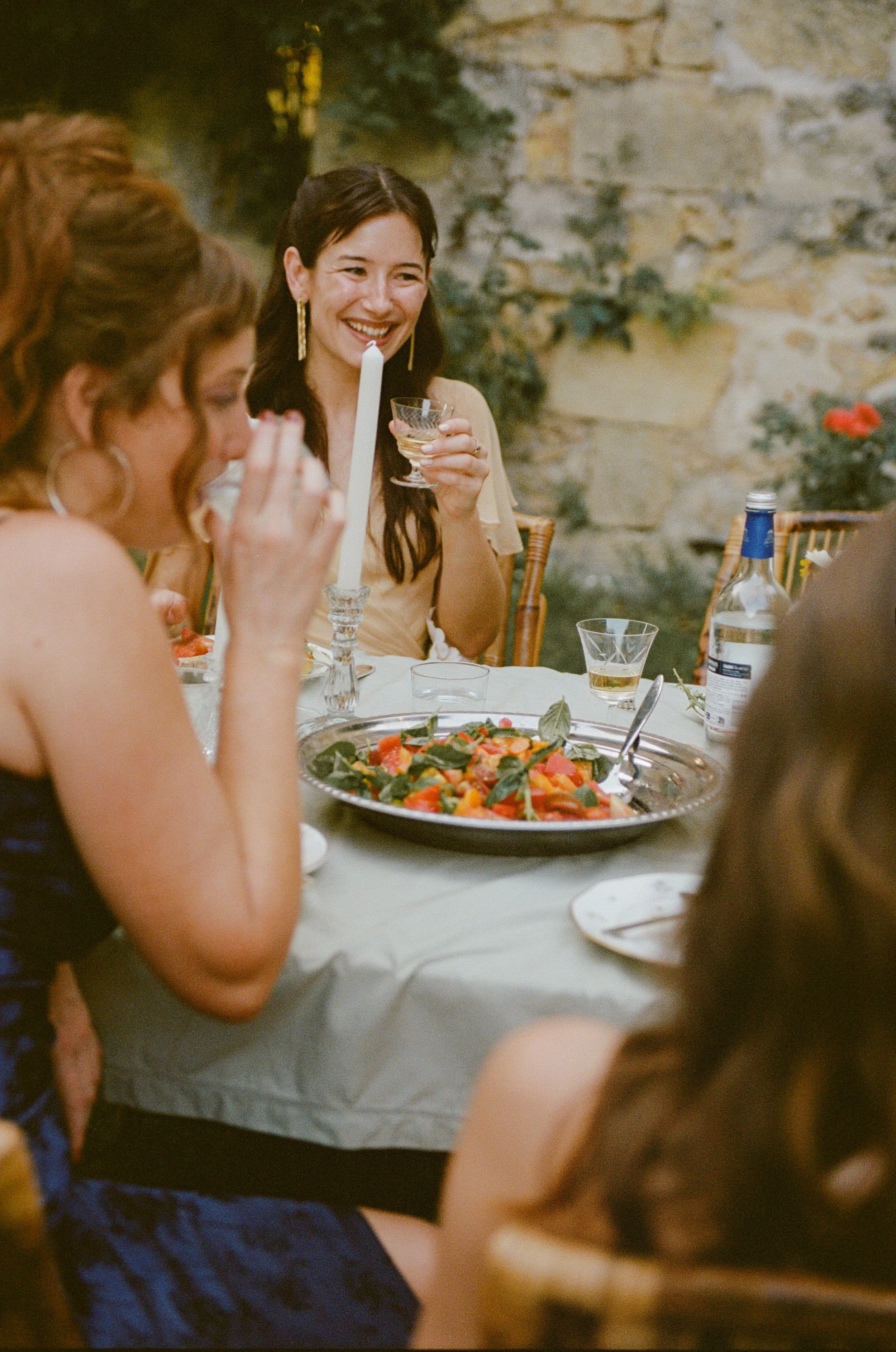 A woman laughing and holding a glass at a dinner party with other women around a table with salad and drinks, outdoor setting with stone wall and greenery in the background.