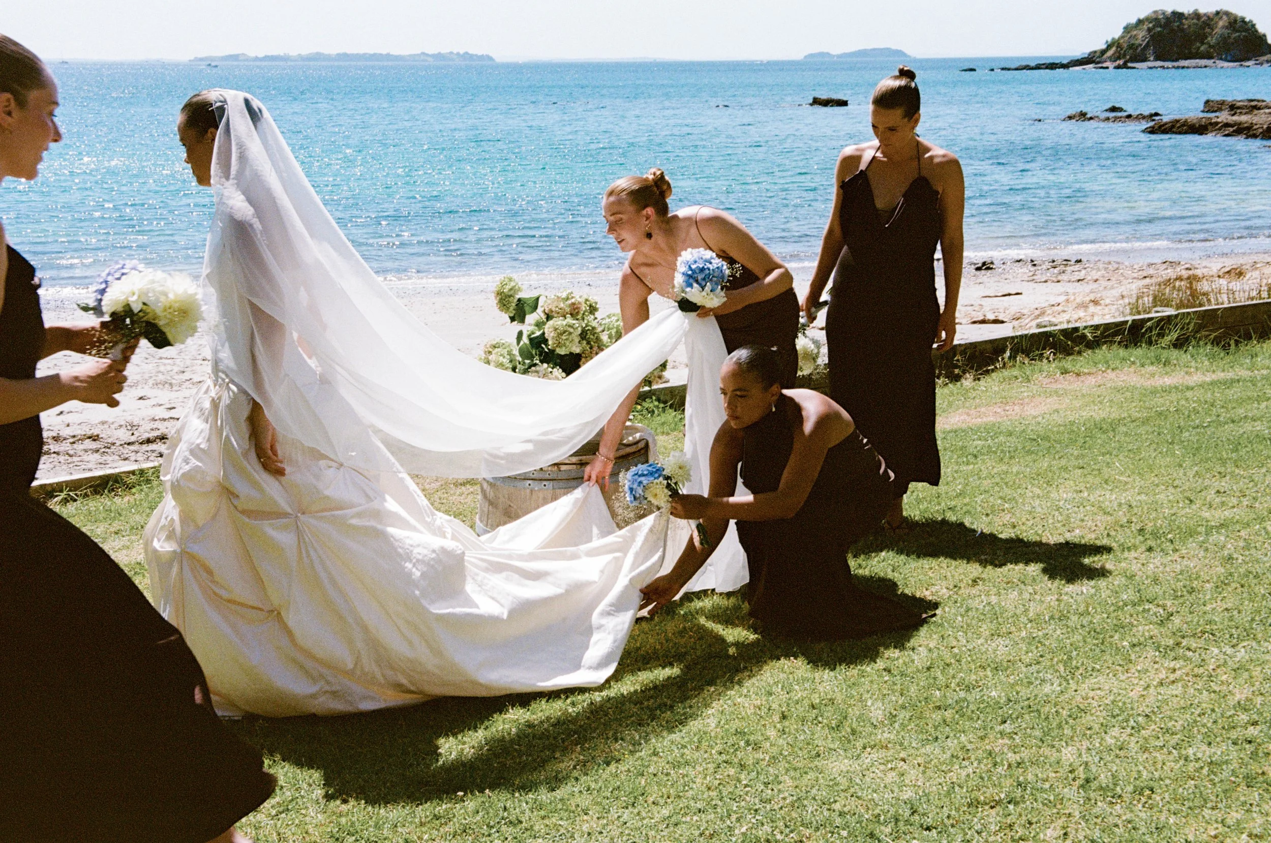 A bride and her bridesmaids on a beach setting, preparing her wedding dress with the ocean in the background.