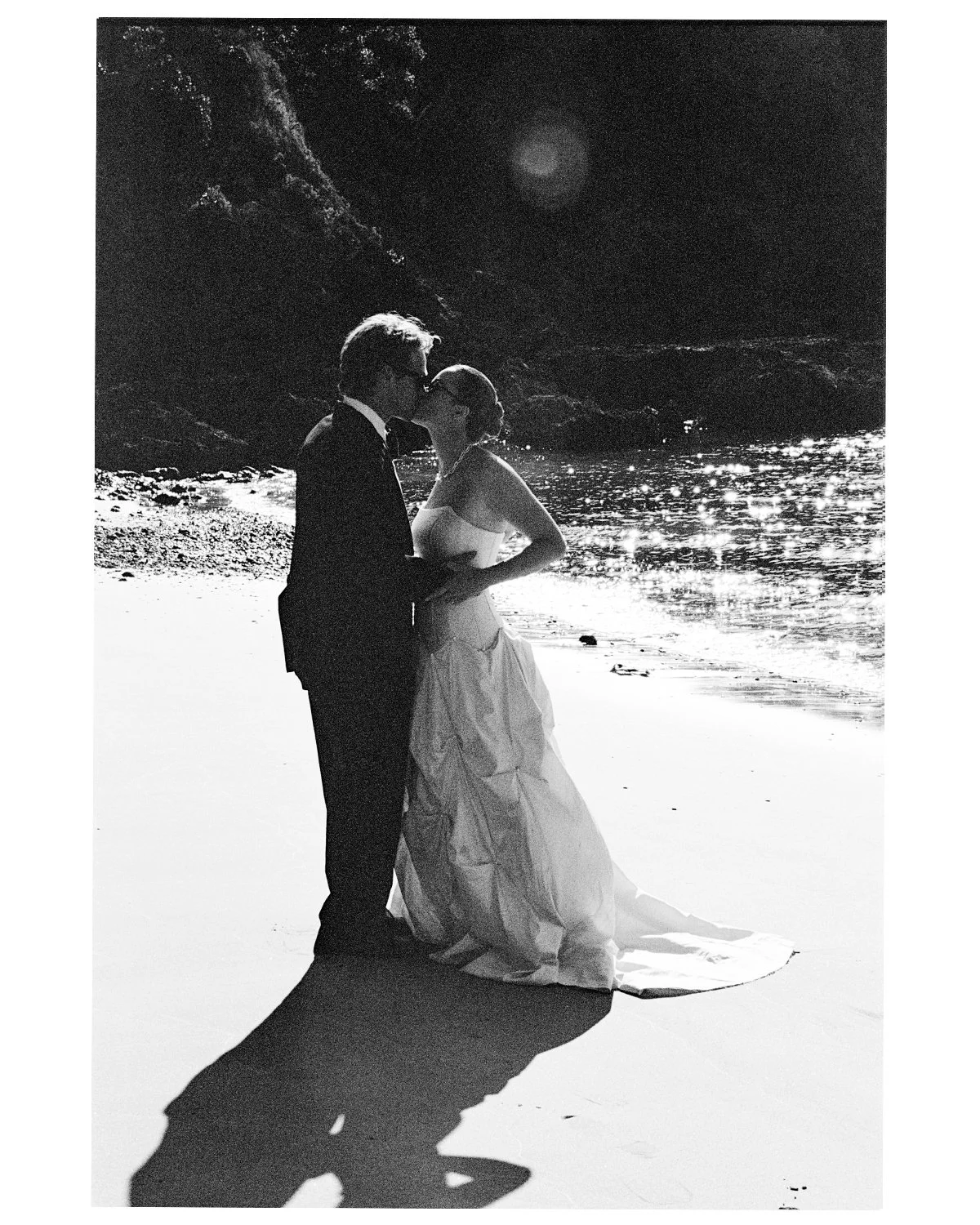 A black and white photo of a couple in wedding attire standing on a beach, sharing a kiss, with water and rocky cliffs in the background.